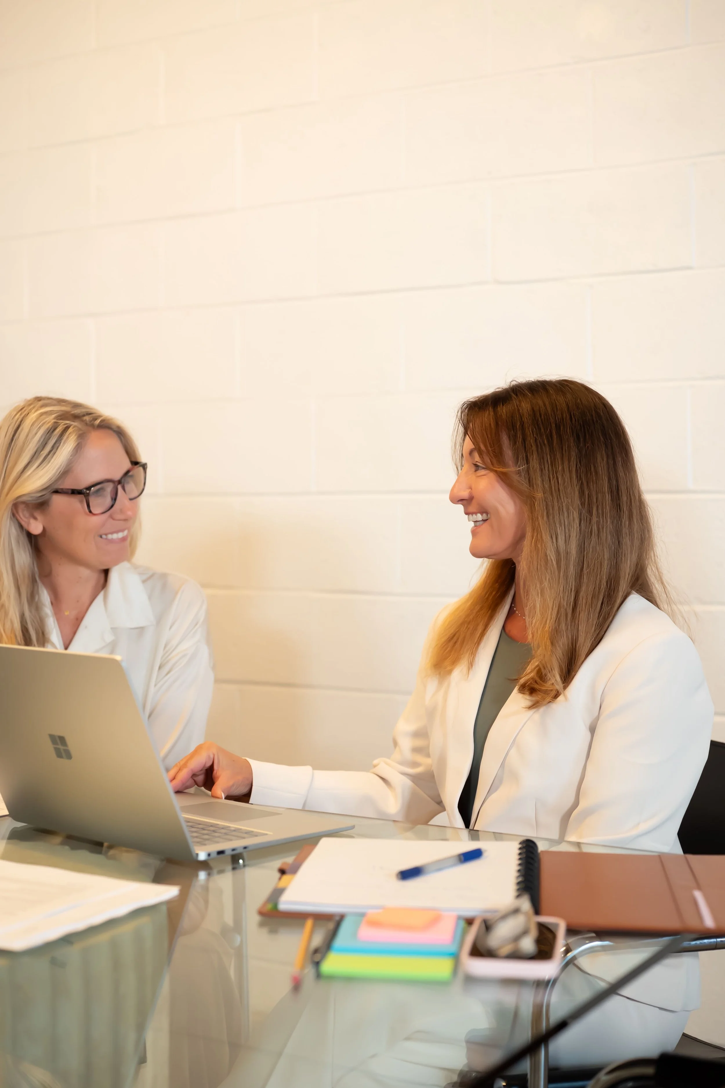Two business women in white, smiling, and working by laptop