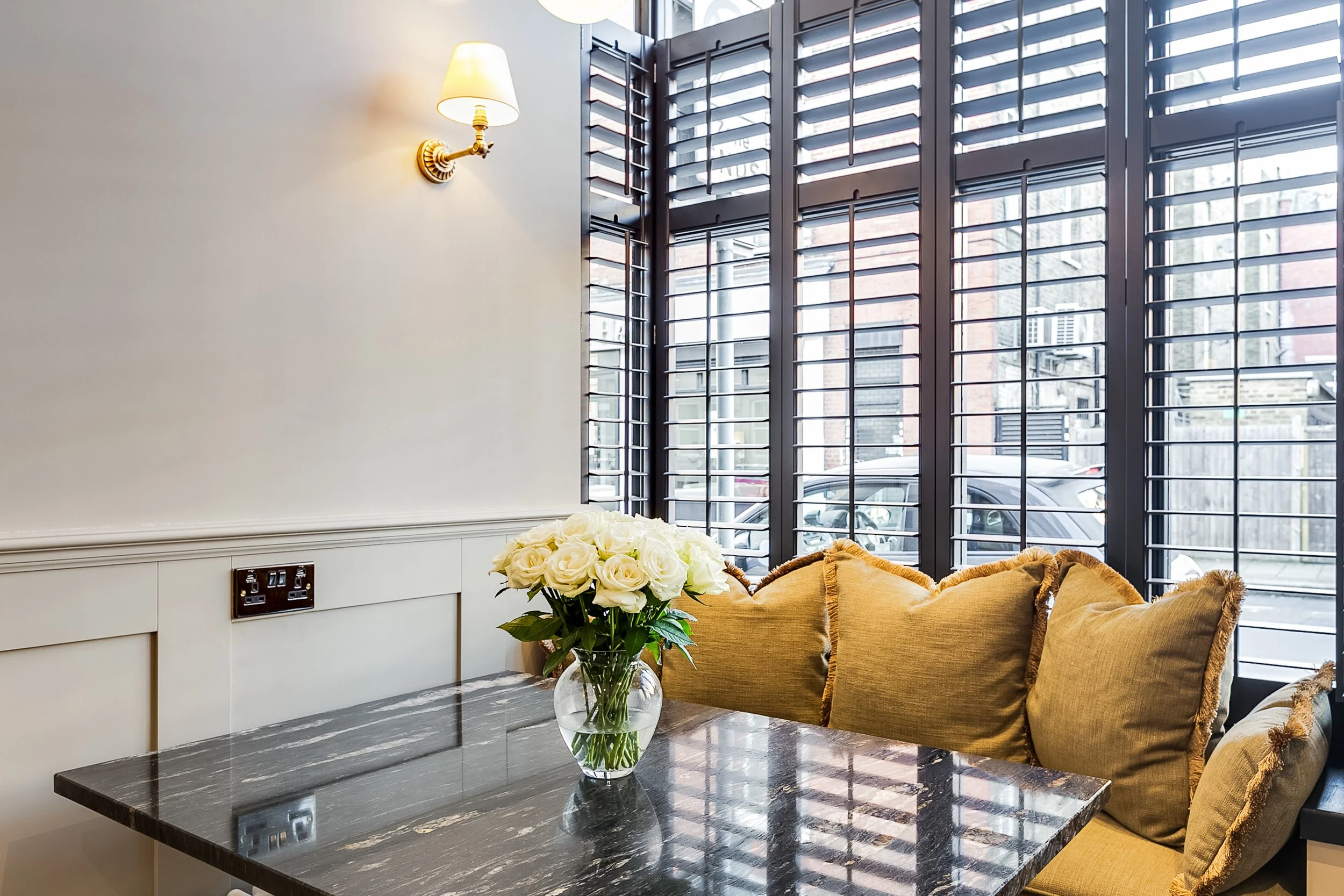 Interior of a room with a window featuring black shutters, a white wall with a wall-mounted lamp, a dark marble table, a bouquet of white roses in a glass vase, and yellow cushions on a sofa.