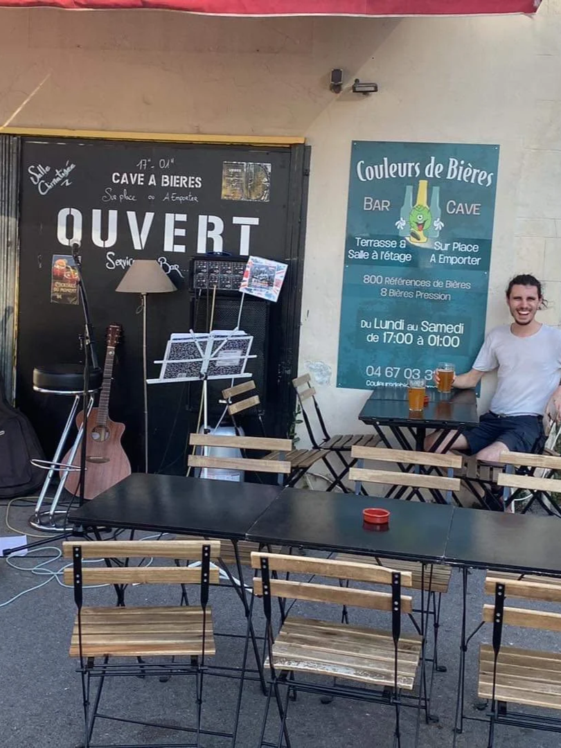 Une terrasse de bar avec des tables en bois et une personne souriante tenant un verre de bière. À gauche, une guitare et un micro. À droite, un panneau bleu avec des informations sur le bar, notamment qu'il est ouvert du lundi au samedi de 17h00 à 01