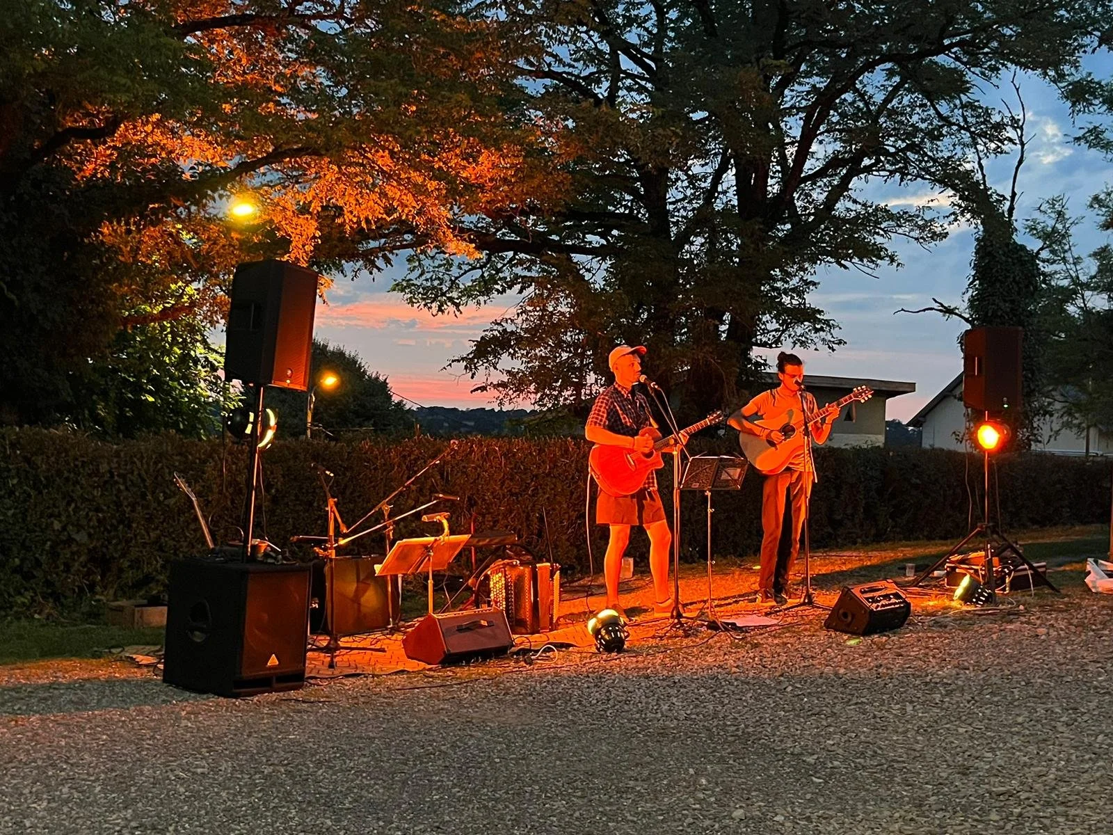 Deux musiciens jouent de la guitare lors d'un concert en plein air au coucher du soleil, entourés d'arbres et de lampes d'ambiance.