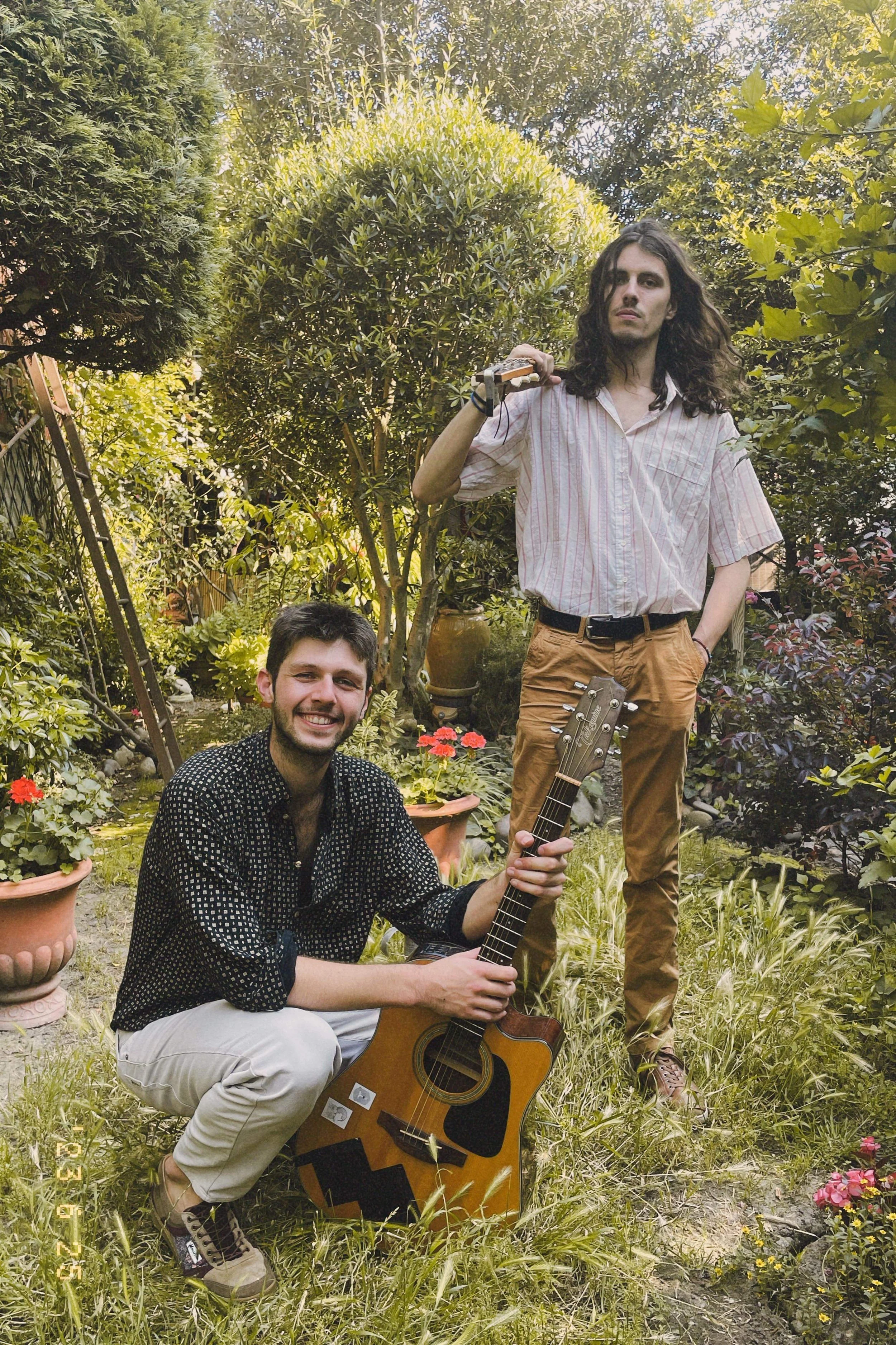 Deux jeunes hommes dans un jardin entouré de plantes et de fleurs. L'un est assis avec une guitare, l'autre se tient debout avec un instrument à la main.