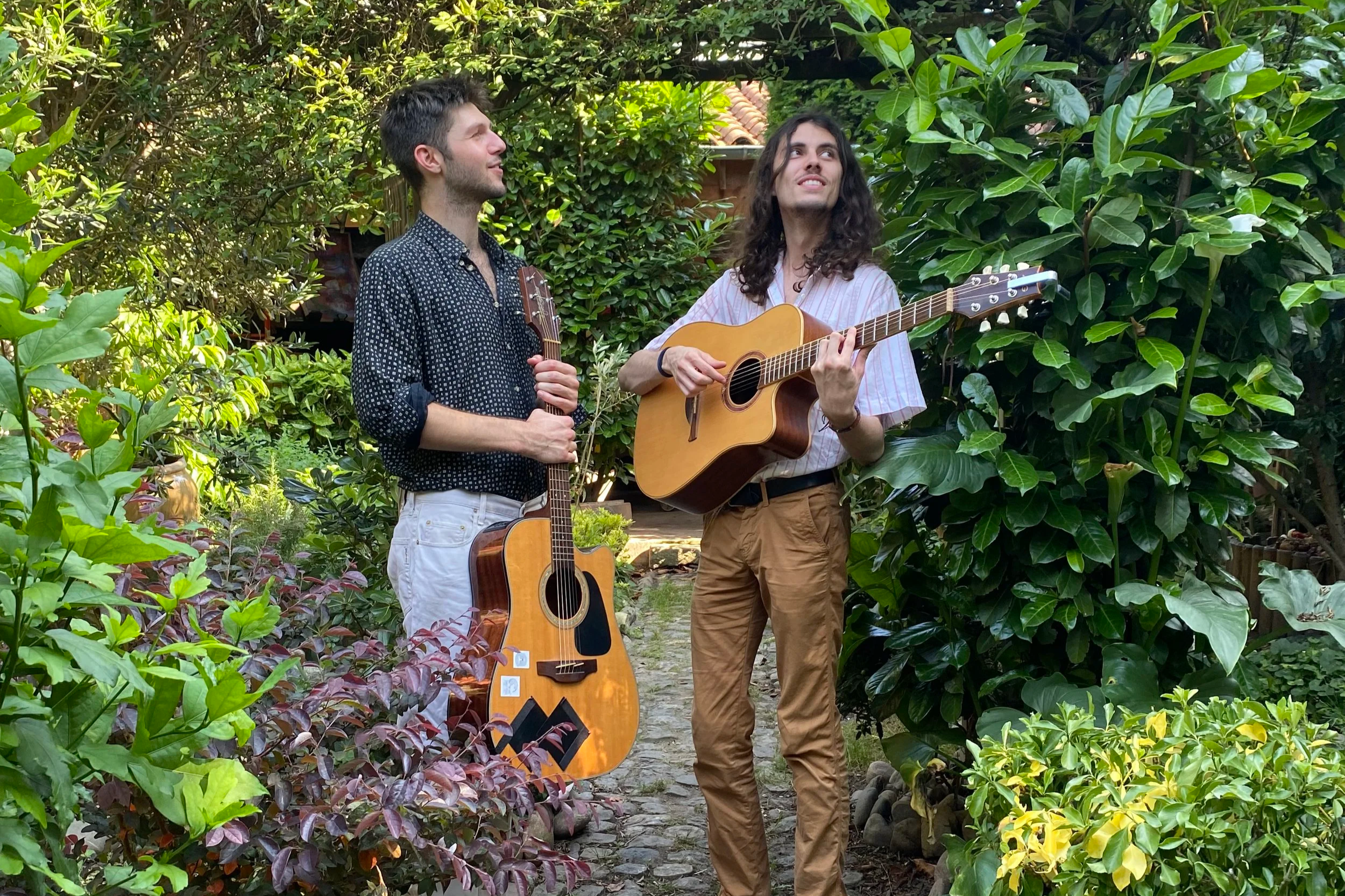 Deux jeunes hommes jouent de la musique en plein air dans un jardin verdoyant. Ils tiennent chacun une guitare.