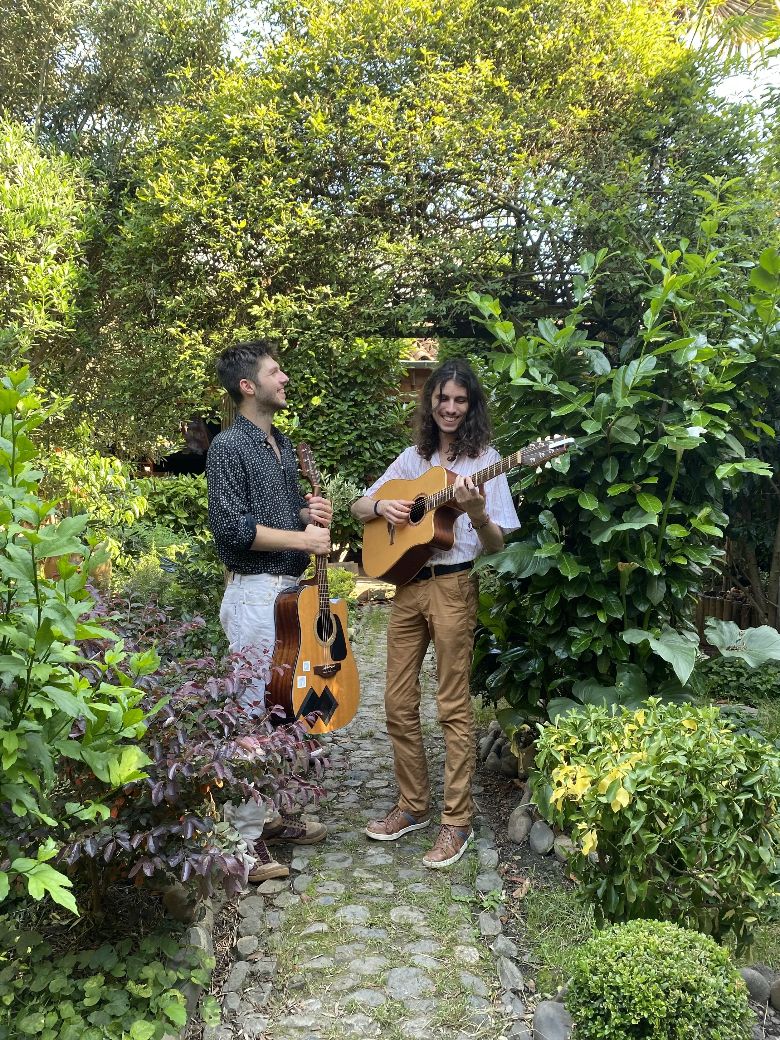 Deux jeunes hommes jouent de la guitare dans un jardin verdoyant, entourés de buissons et d'arbres, en plein jour.
