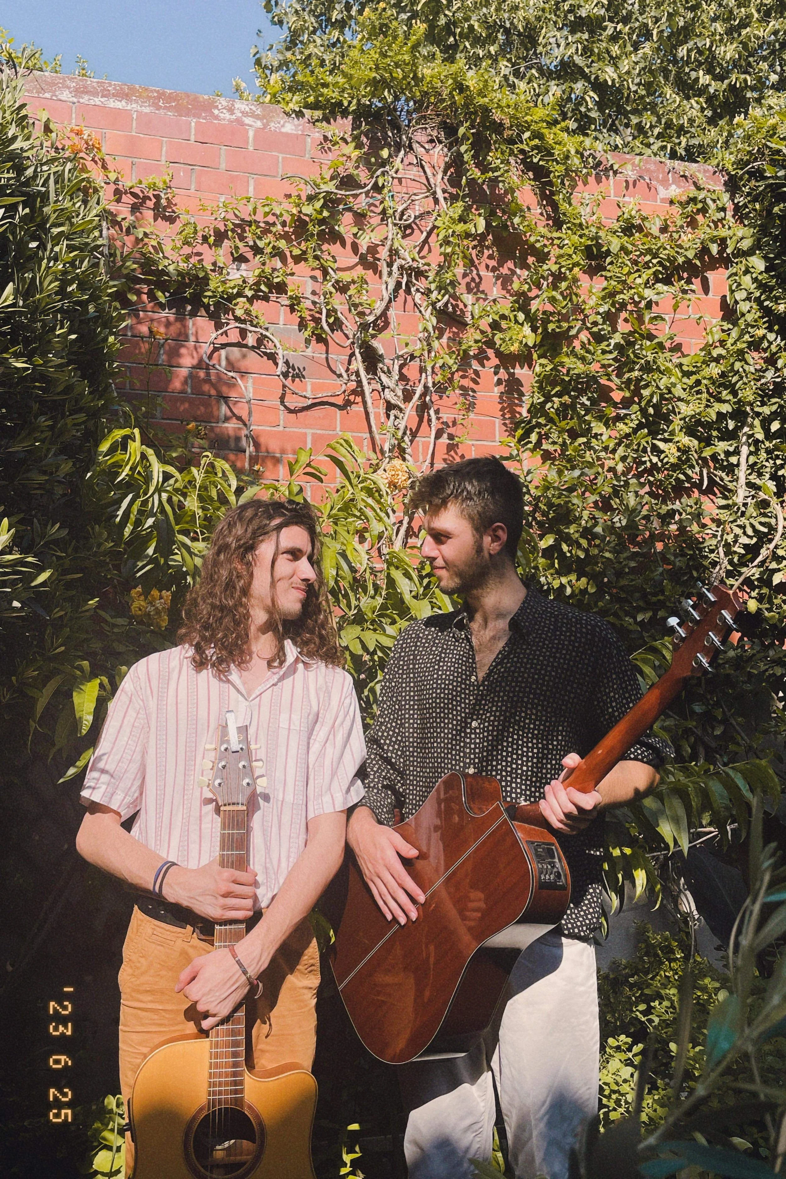 Deux jeunes hommes chantant ou jouant de la musique dans un jardin, avec une marqueterie de plantes et un mur en briques rouges en arrière-plan, en plein jour.