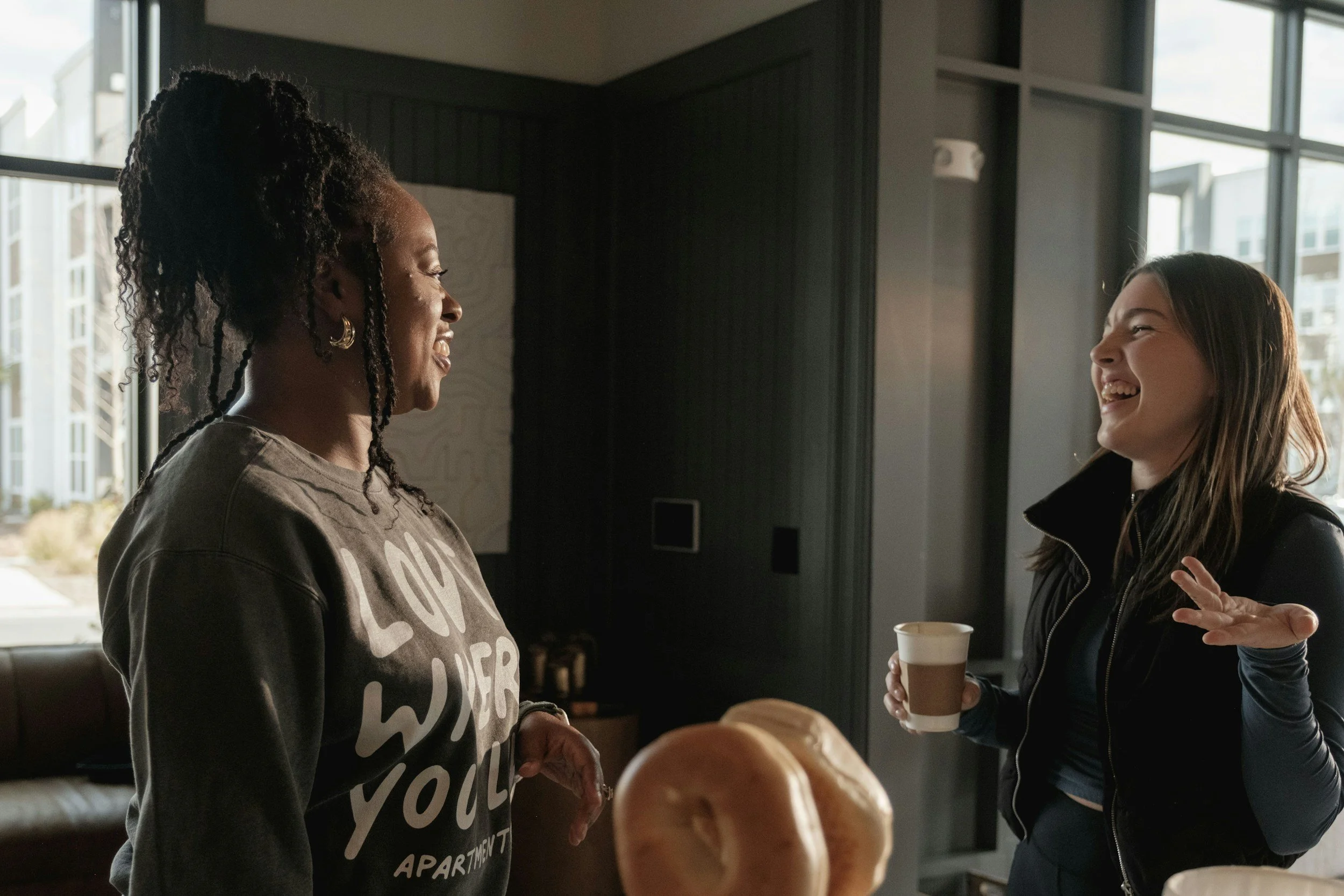 Two women smiling and talking in a warmly lit room with large windows, one holding a coffee cup.