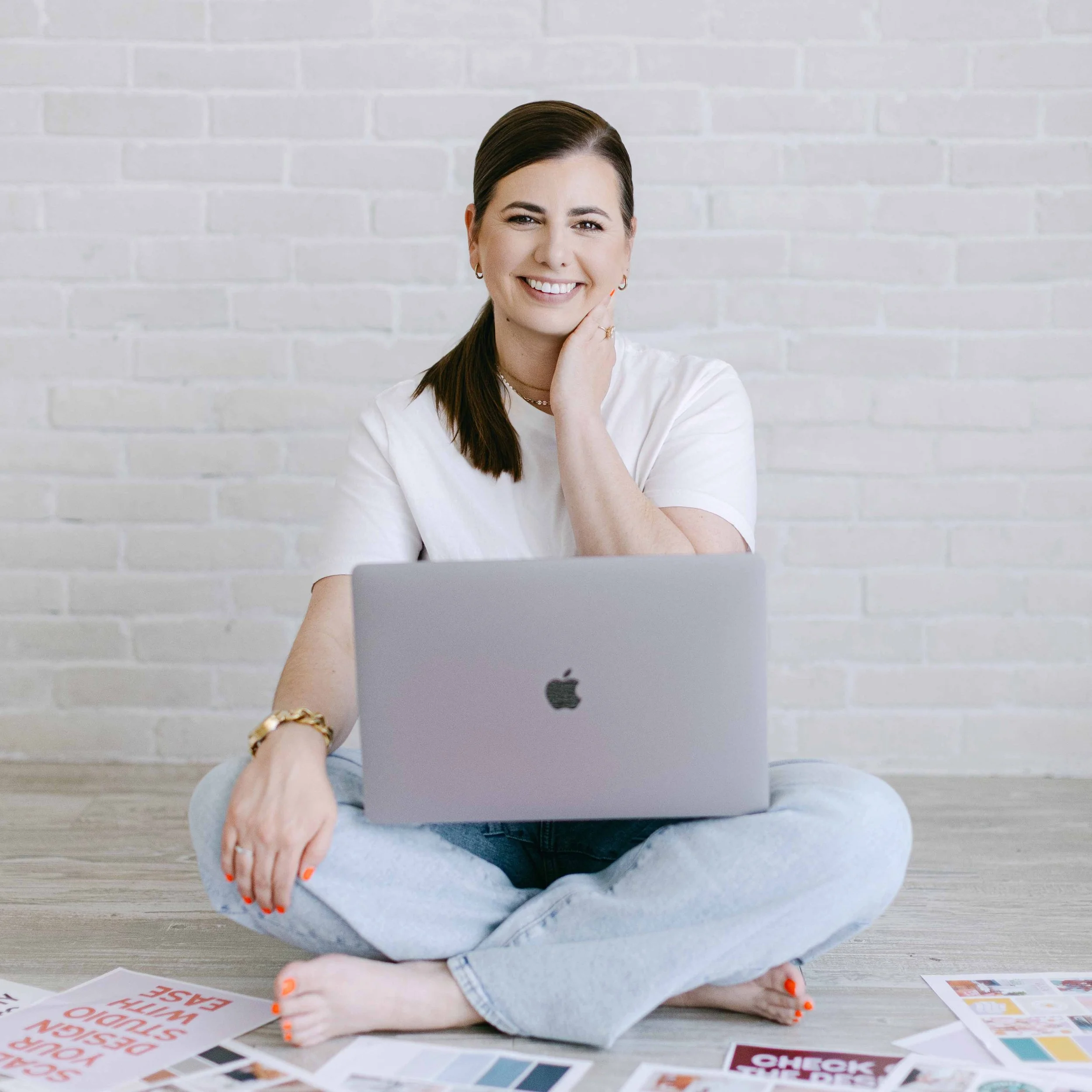 Woman sitting on the floor with laptop, looking at the camera, smiling, surrounded by colorful flyers on the floor.
