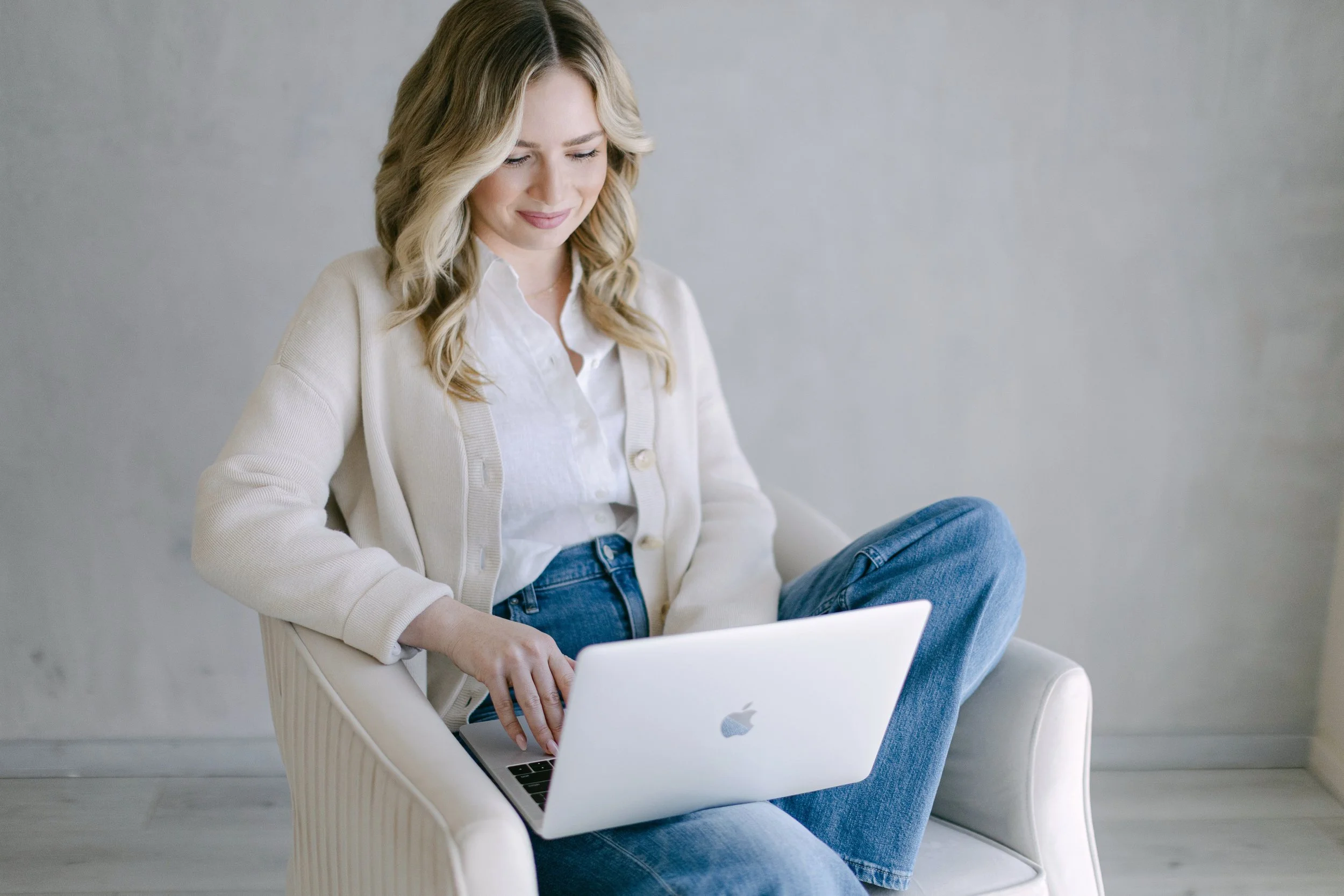A young woman with wavy blonde hair, wearing a white button-up shirt, a cream cardigan, and blue jeans, sitting on a white chair and using a silver MacBook laptop.