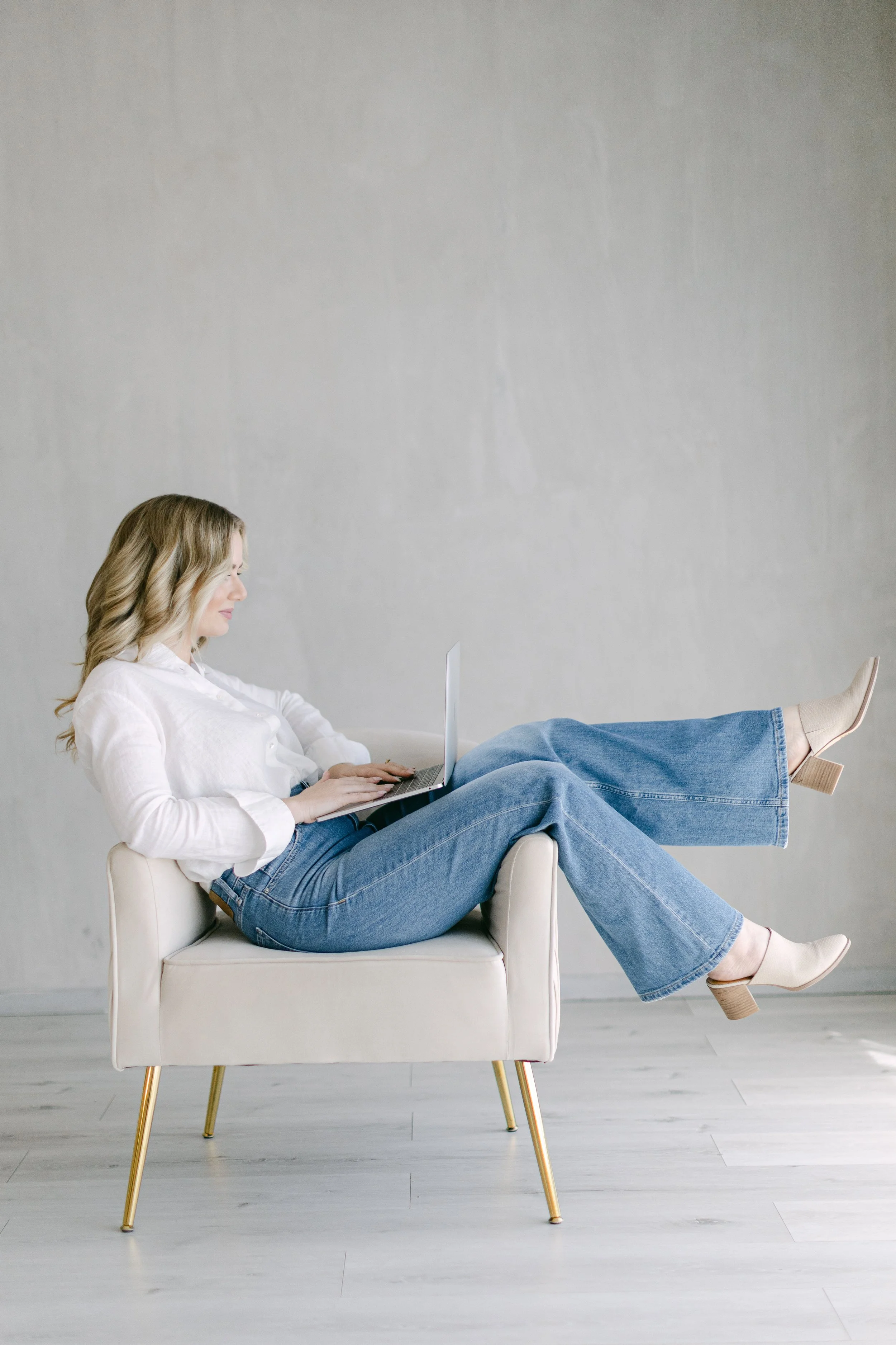 A woman with blonde hair sitting on a cream-colored armchair, using a laptop, with her legs resting on the armrest and feet in beige heels, against a plain gray wall.