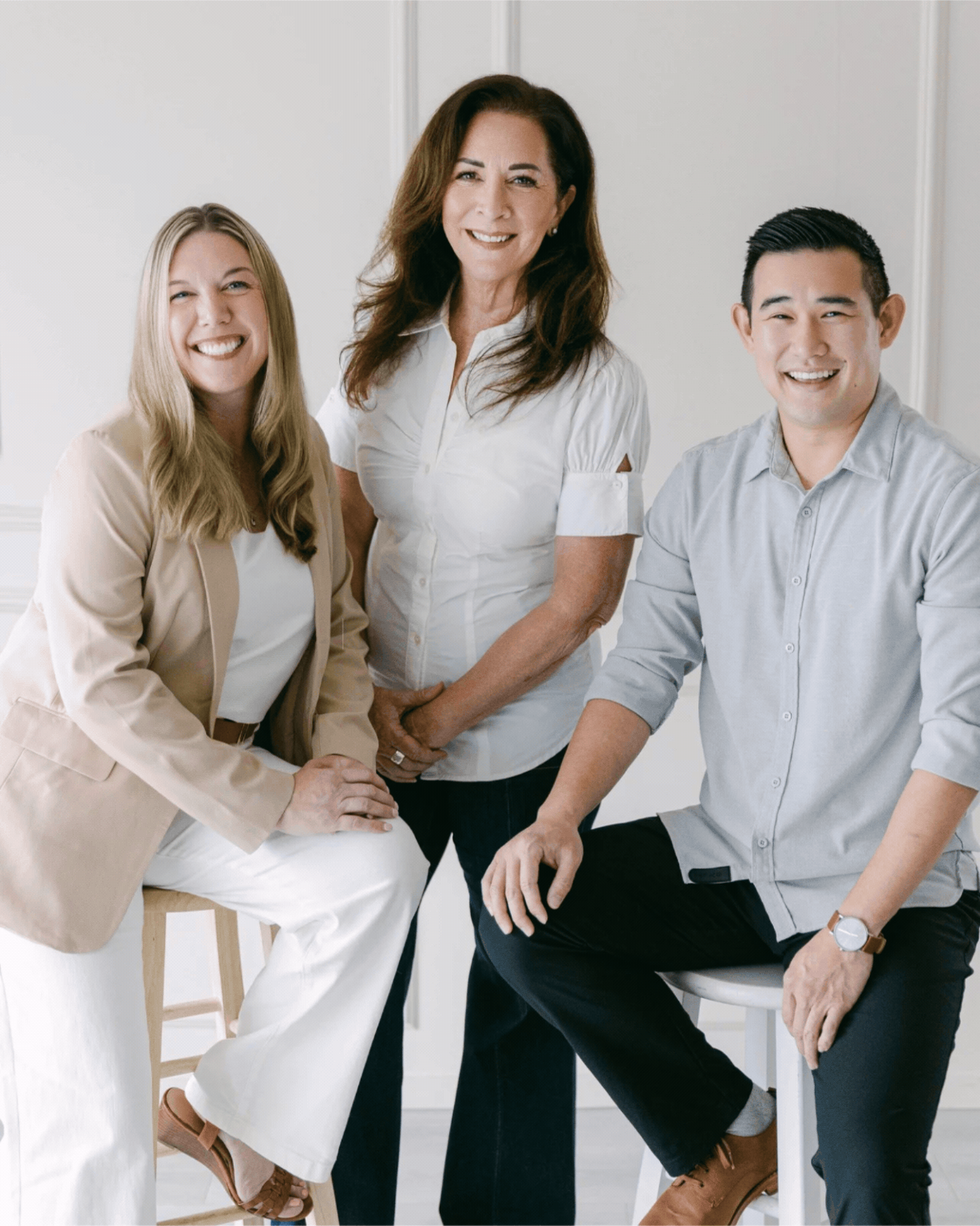Three smiling people, two women and one man, dressed in business casual, posing in a bright room with a white background.