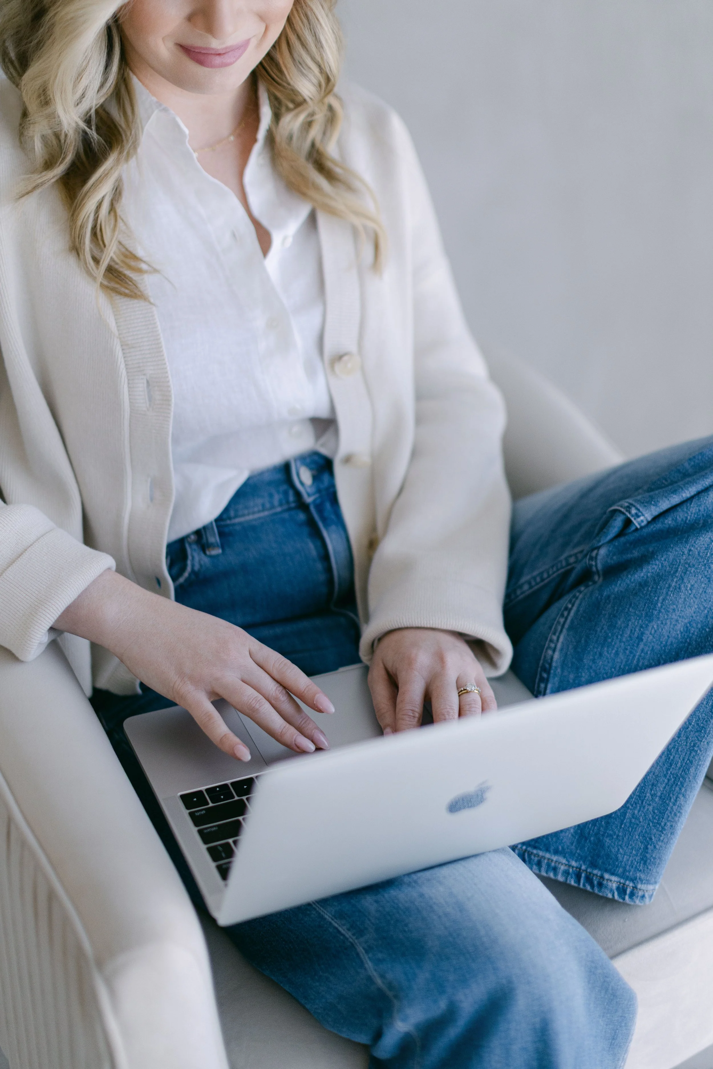 A woman with blonde hair wearing a white shirt and beige cardigan sitting on a beige chair, using a silver laptop with a black keyboard placed on her lap.