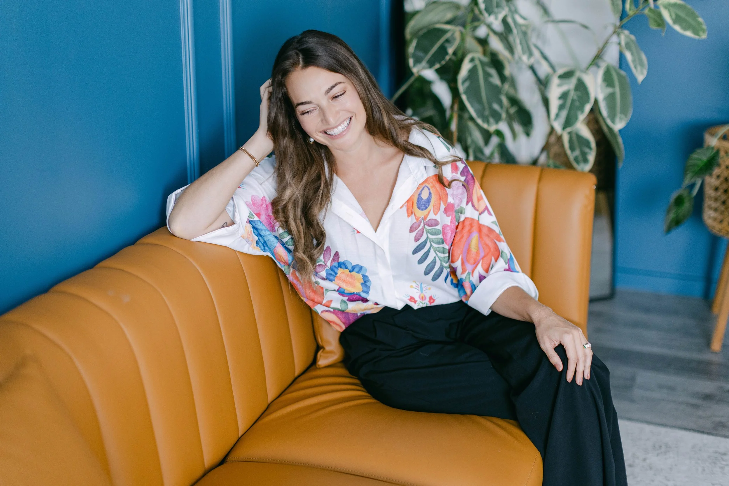 A woman with long brown hair, smiling, sitting on an orange leather sofa. She is wearing a white shirt with colorful floral patterns and black pants. She has her left hand resting on her head, and she appears to be happy. In the background, there is a large leafy green plant and a blue wall.