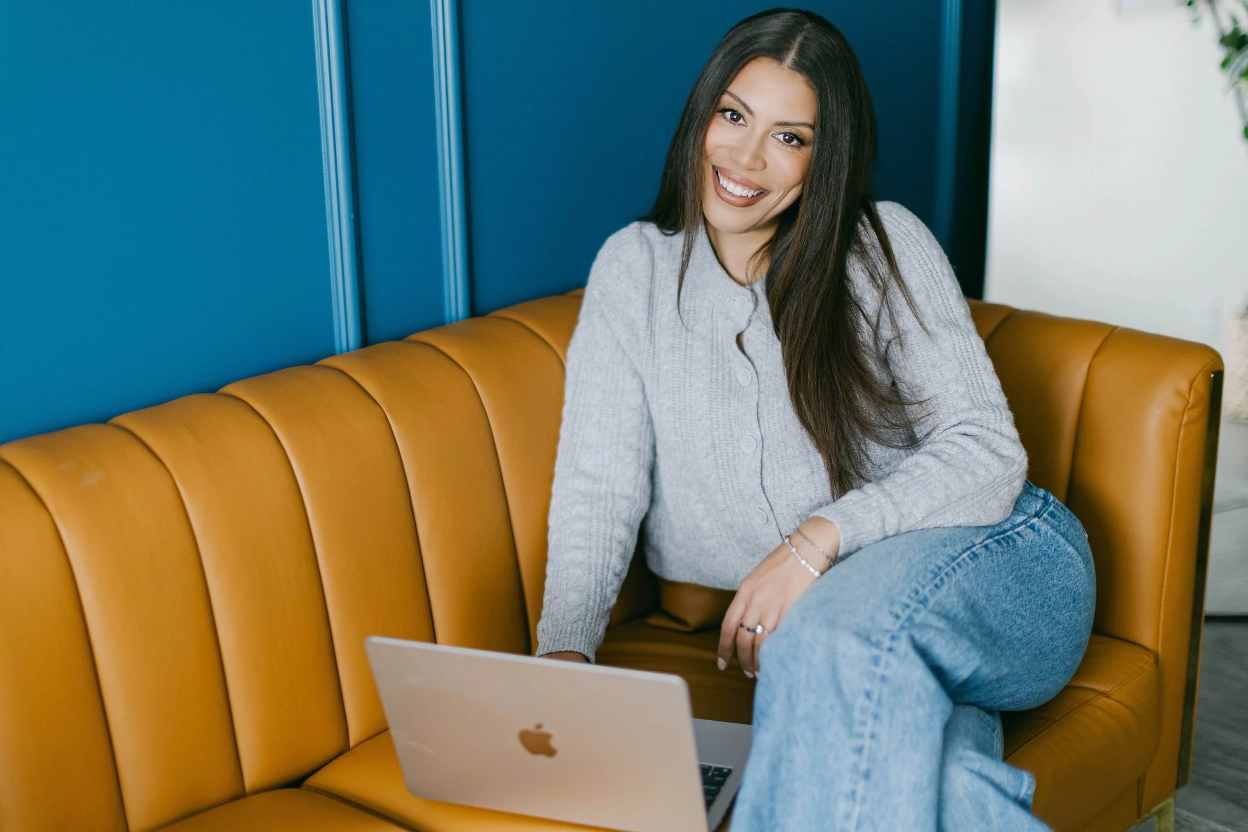 A young woman with long dark hair sitting on a yellow leather couch, smiling at the camera, with a silver laptop in front of her and a blue wall behind her.