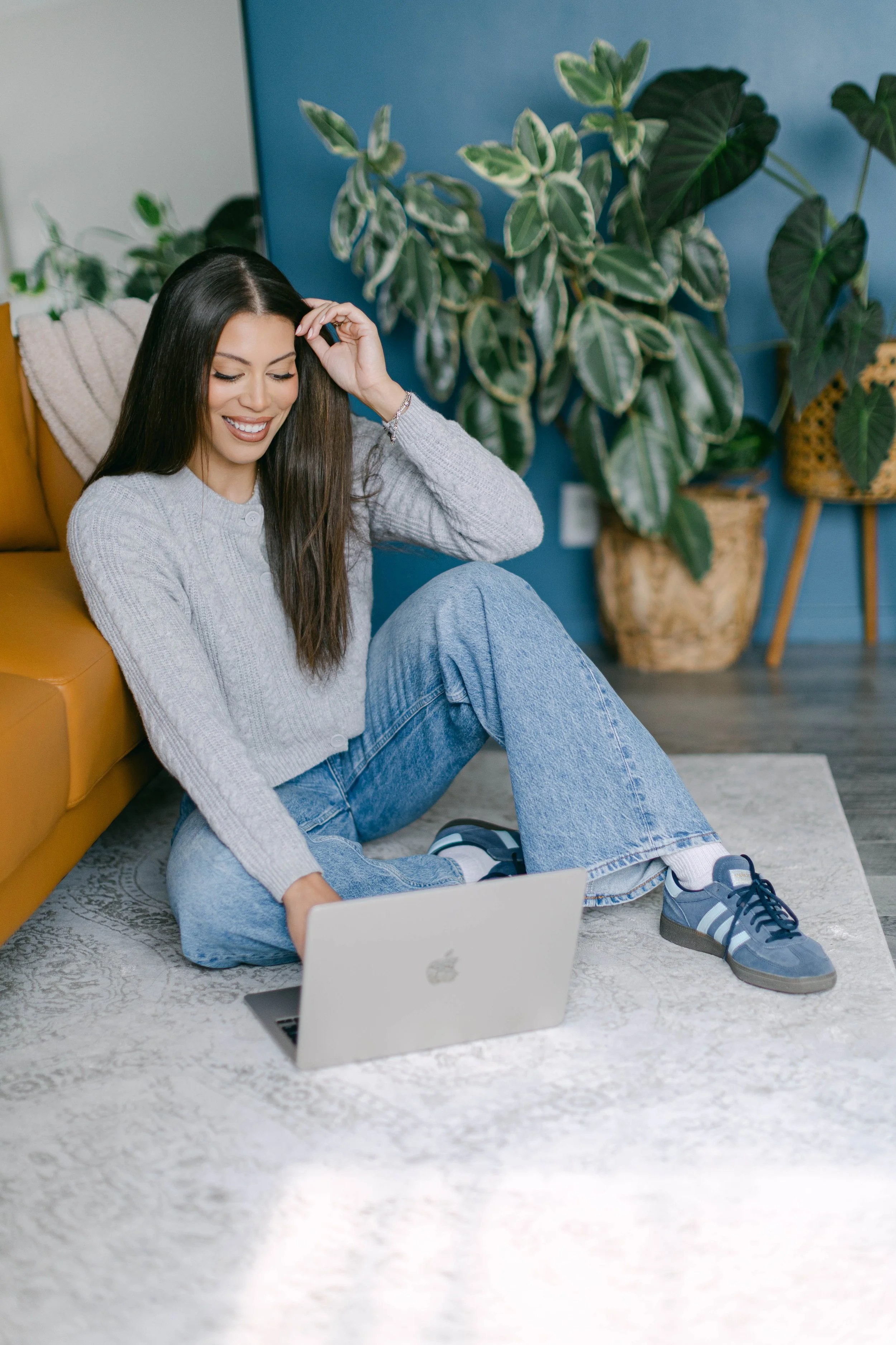 A young woman with long dark hair, sitting on a light-colored rug, smiling and looking down at her laptop on the floor. She's wearing a gray button-up sweater, blue jeans, and blue sneakers. Behind her is a mustard-colored couch, large leafy houseplants in woven baskets, and a blue wall.