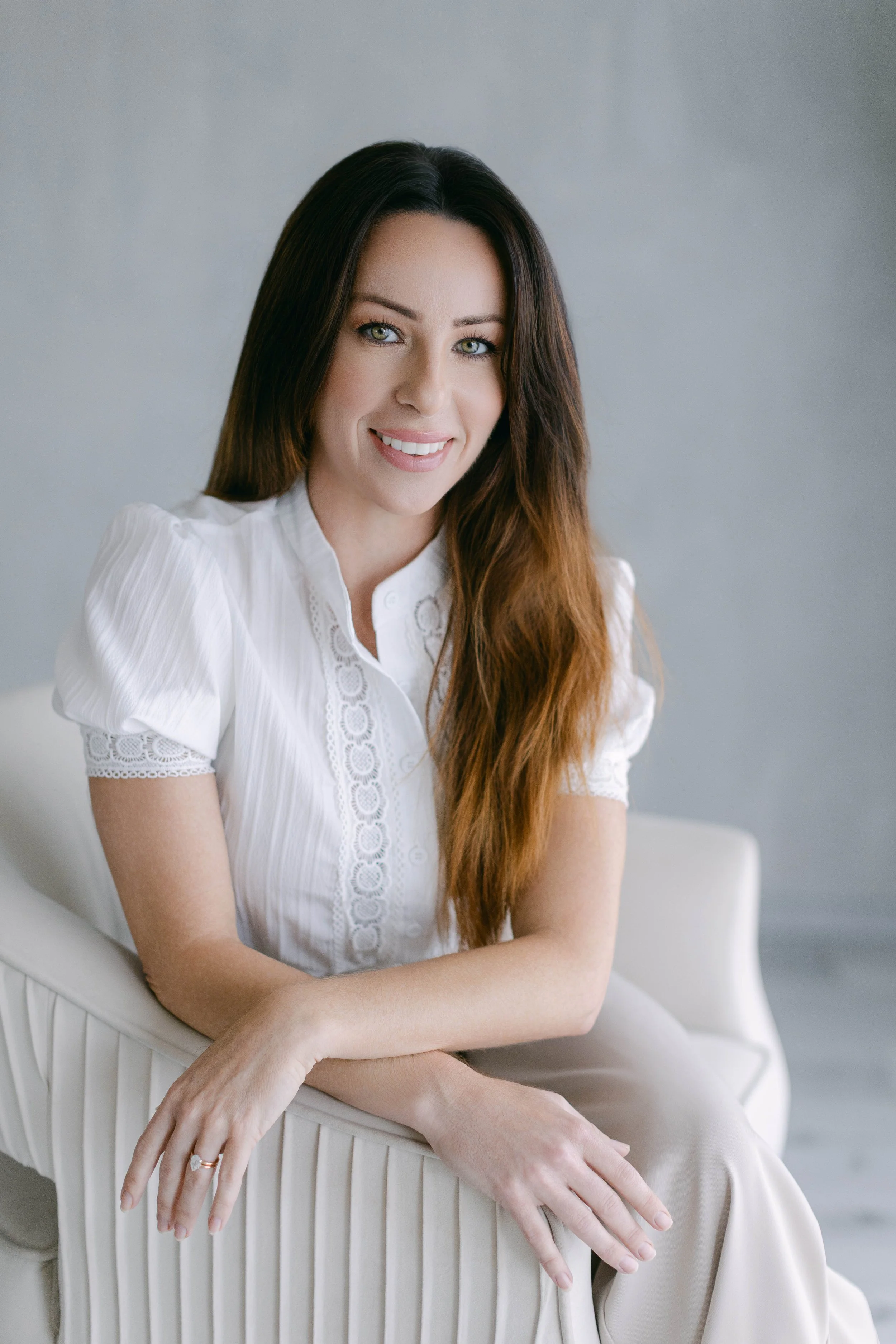 A woman with long brown hair and green eyes, wearing a white blouse, sitting on a white chair with vertical ridges, smiling at the camera against a plain, light gray background.
