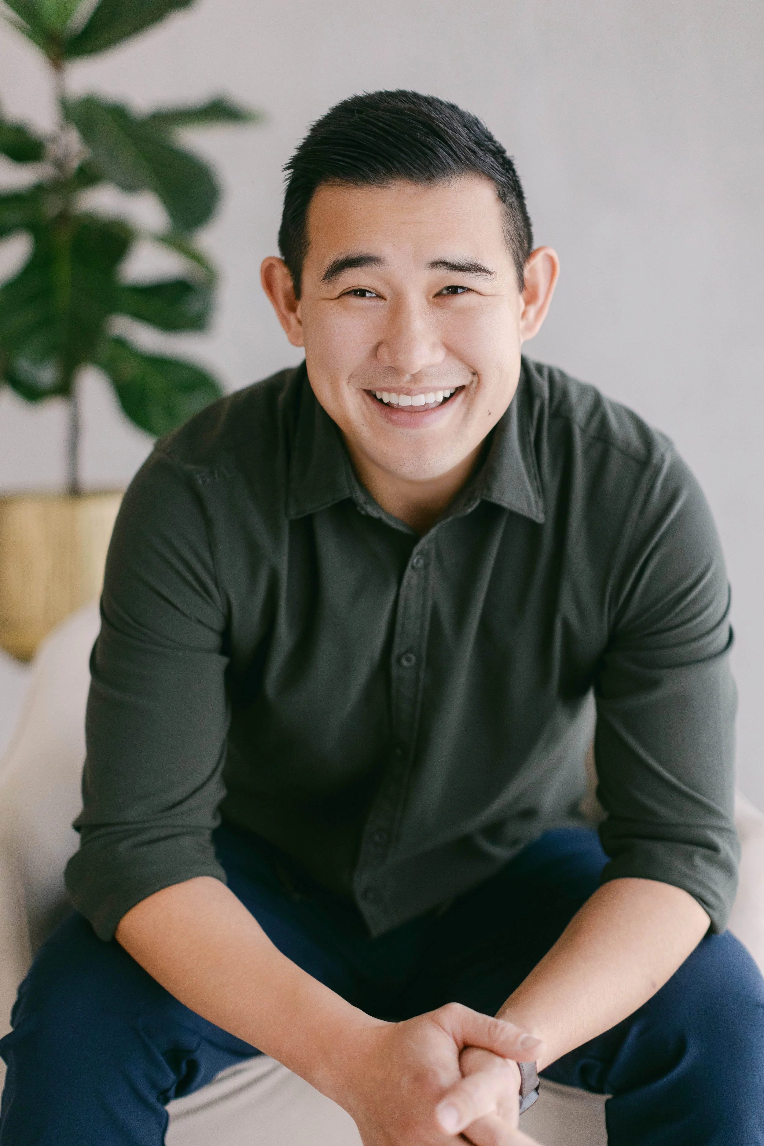 A smiling man with short black hair wearing a dark gray shirt, sitting on a light-colored couch with a plant in the background.