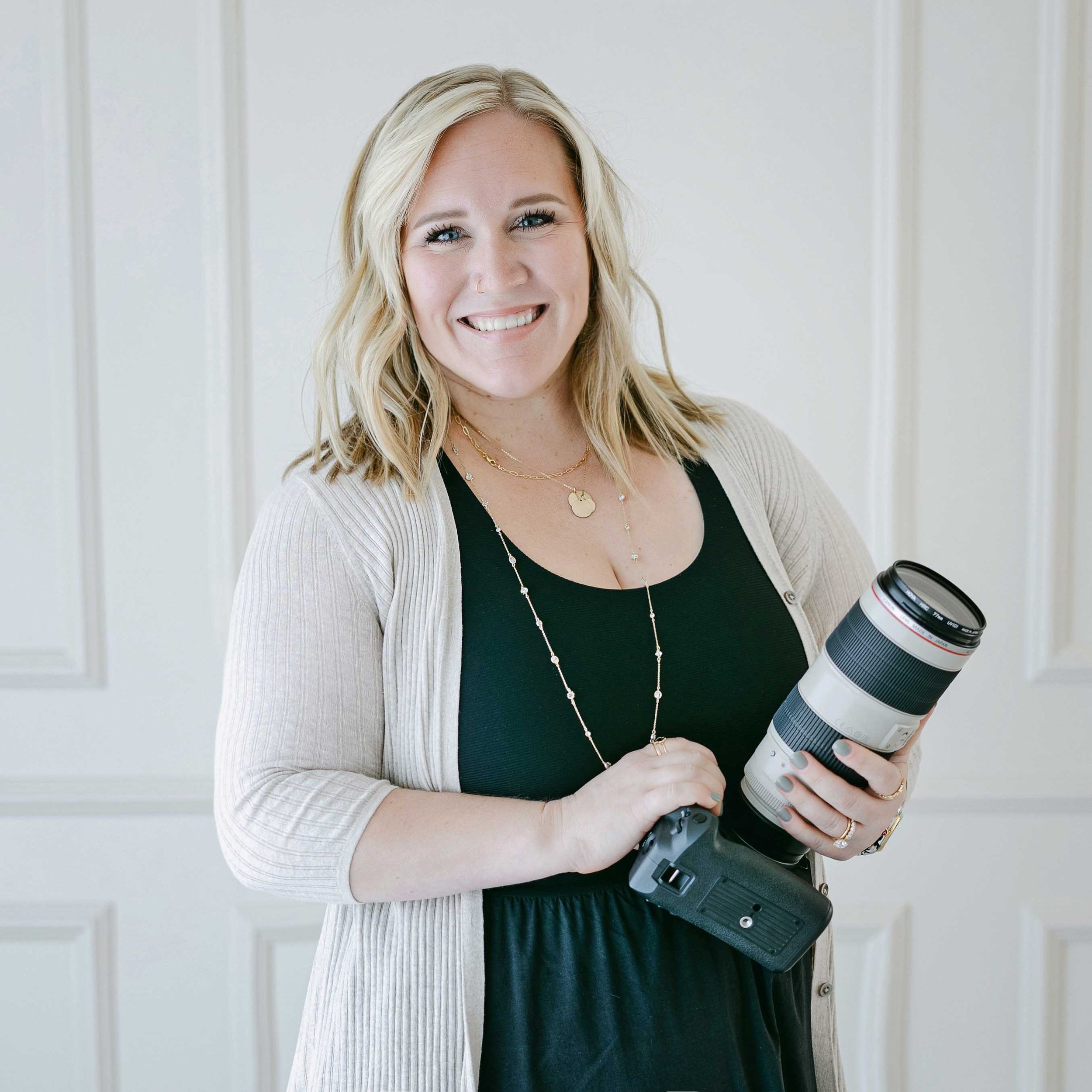 A smiling blonde woman holding a large camera lens in her hand, dressed in black and beige, standing against a white wall with vertical paneling.