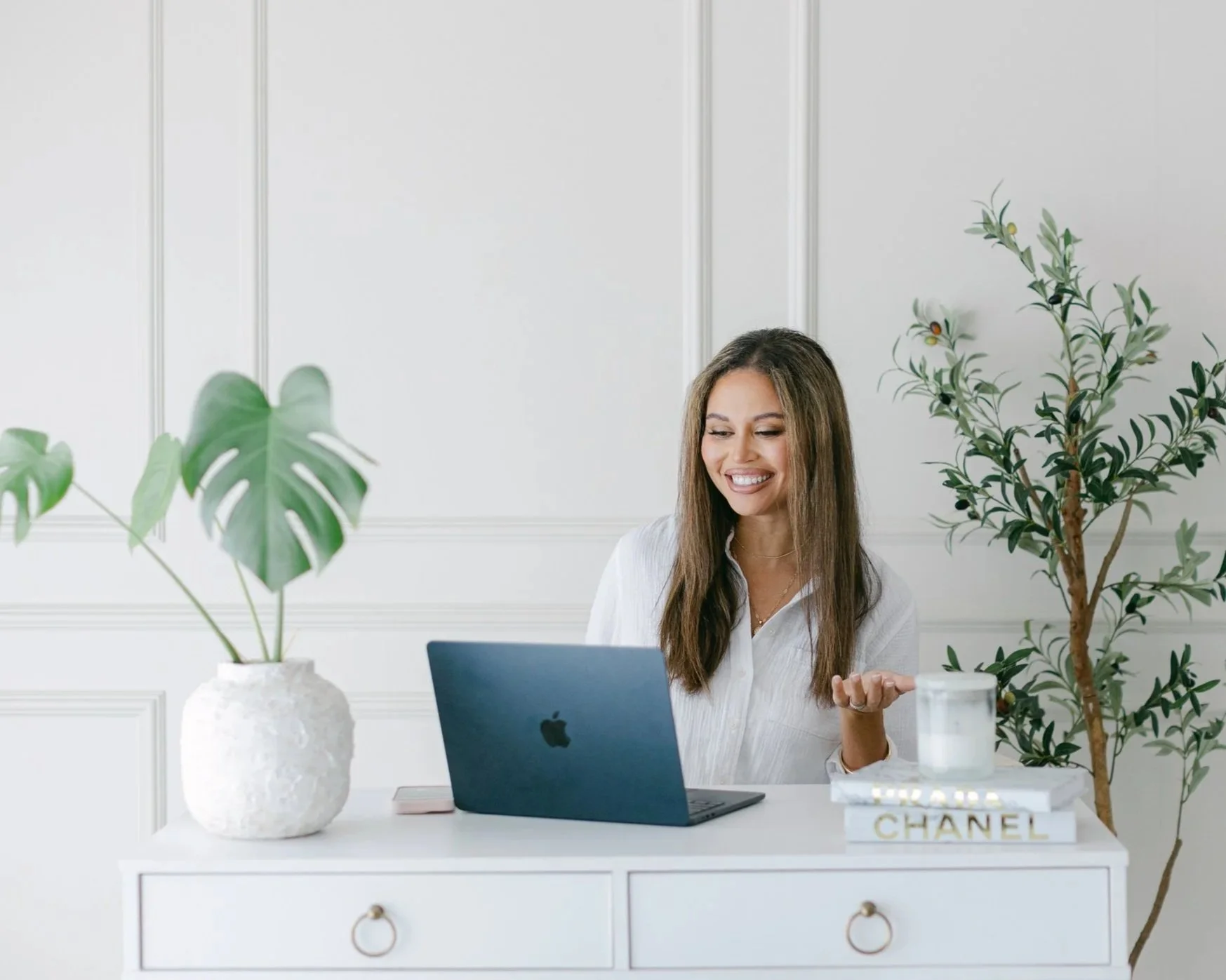 A woman sitting at a white desk with plants, a laptop, and a Chanel book, smiling and looking at her watch.