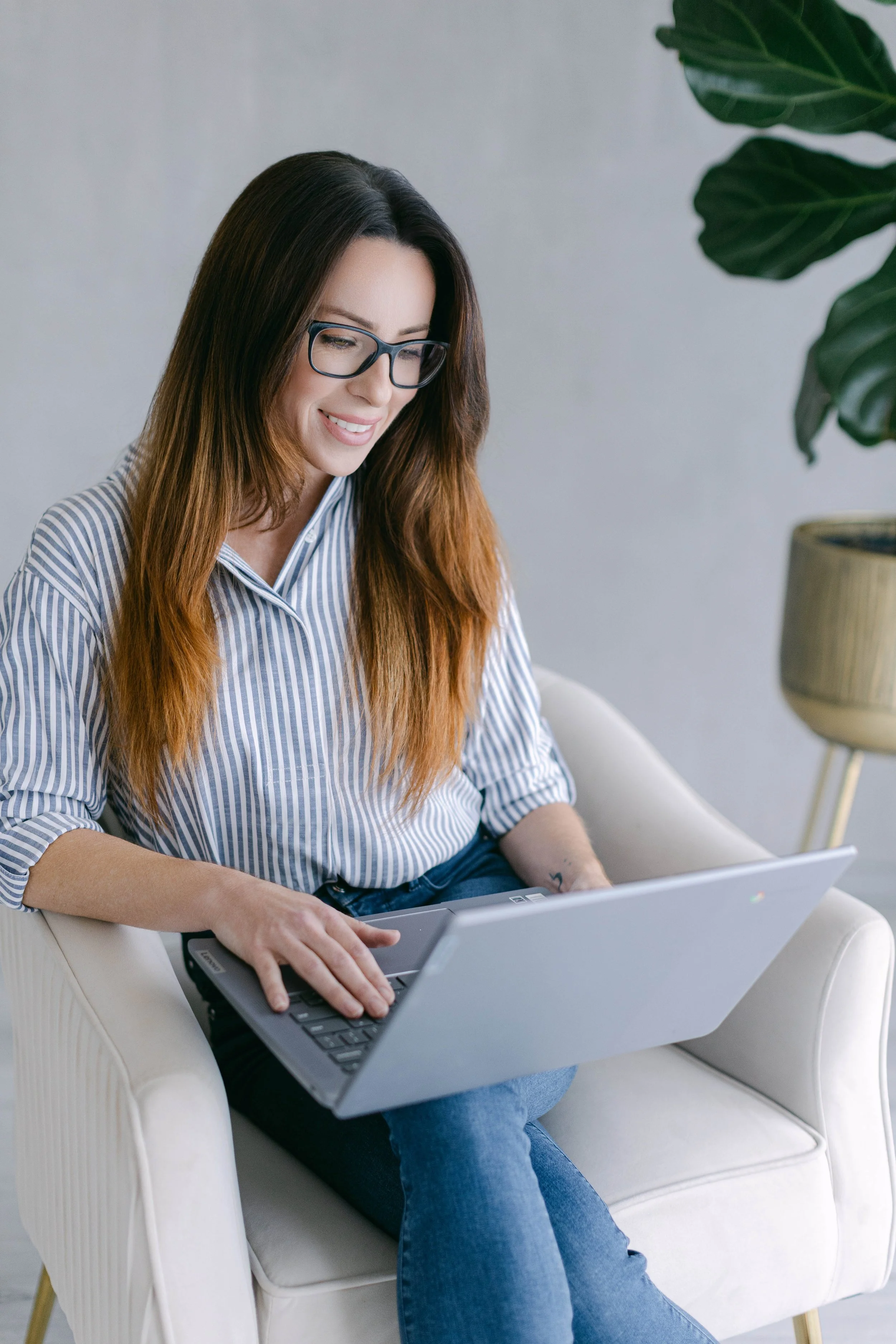 A young woman with long brownish hair, wearing glasses and a blue and white striped shirt, is sitting on a white armchair, working on a laptop, with a large green leafy plant in the background.