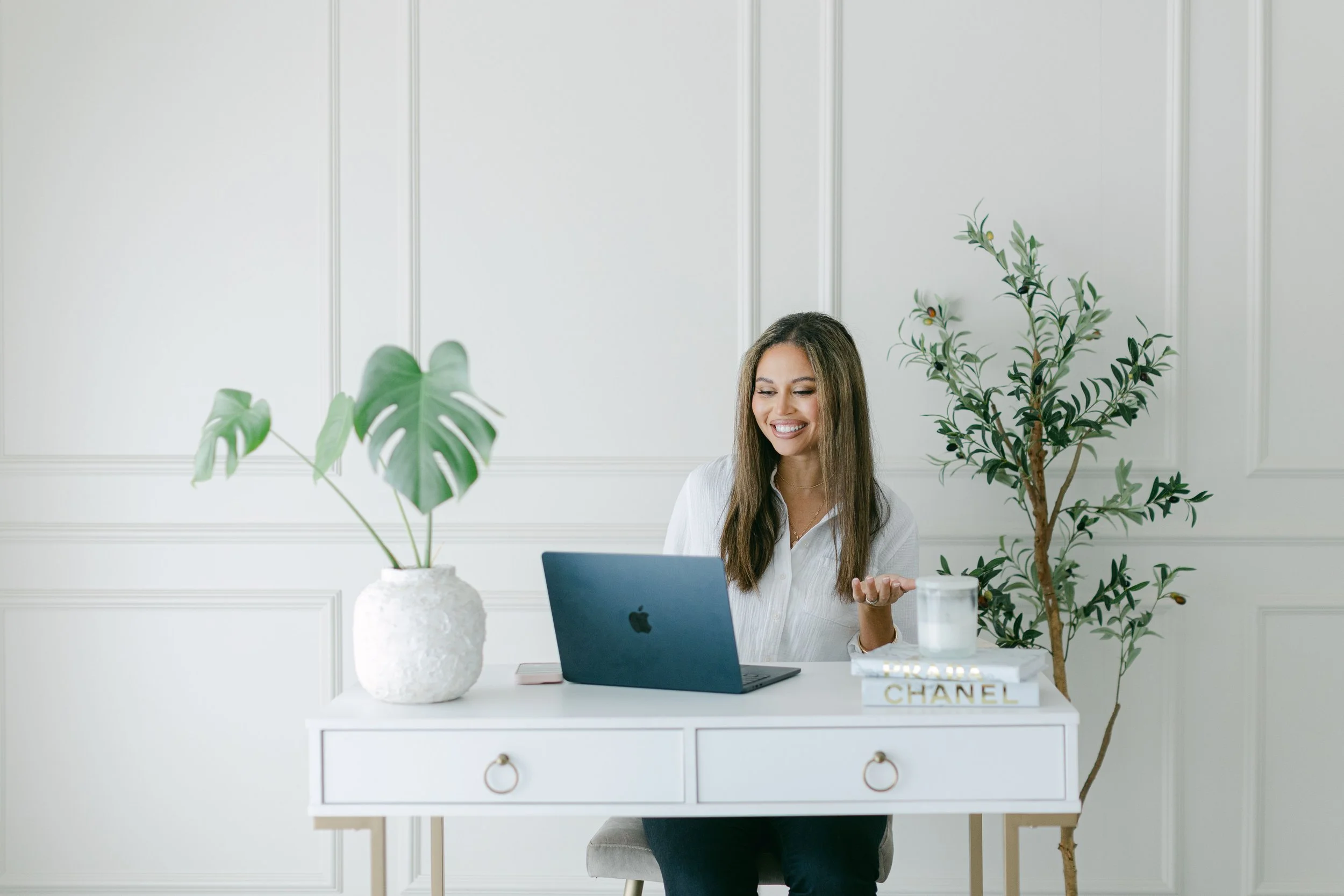 A woman with long brown hair sitting at a white desk, smiling and looking at her laptop, surrounded by green houseplants in a bright, minimalist room.
