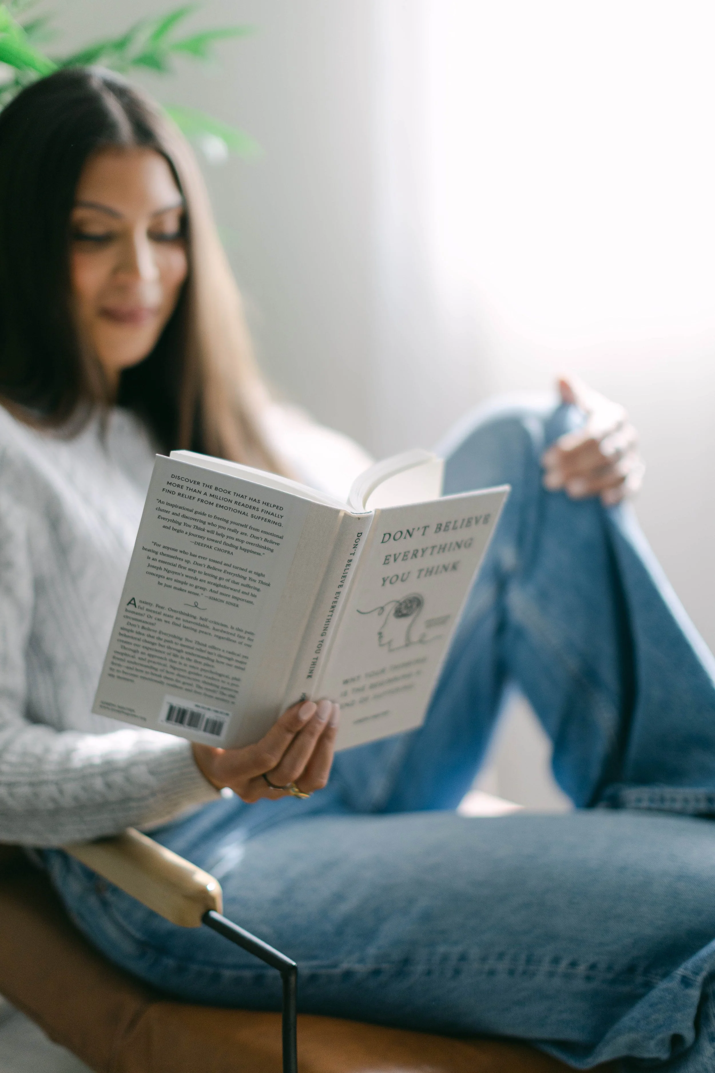 A woman with long brown hair and light skin is sitting in a chair, reading a book titled 'Don't Believe Everything You Think.' She is wearing a white sweater and blue jeans, and is in a well-lit room with a plant in the background.