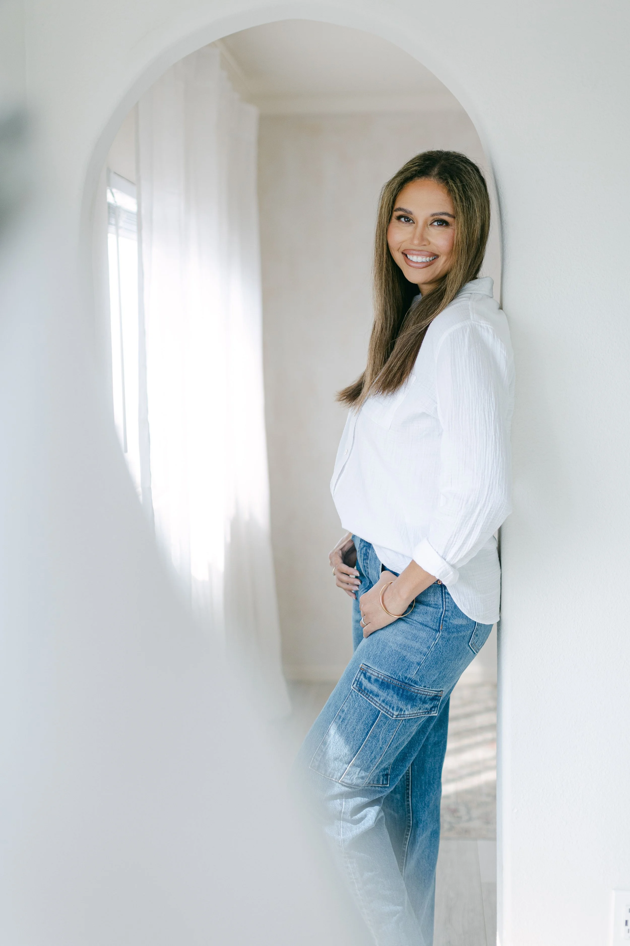 A woman with long hair, smiling and wearing a white shirt and blue jeans, leaning against a white wall in a well-lit room.