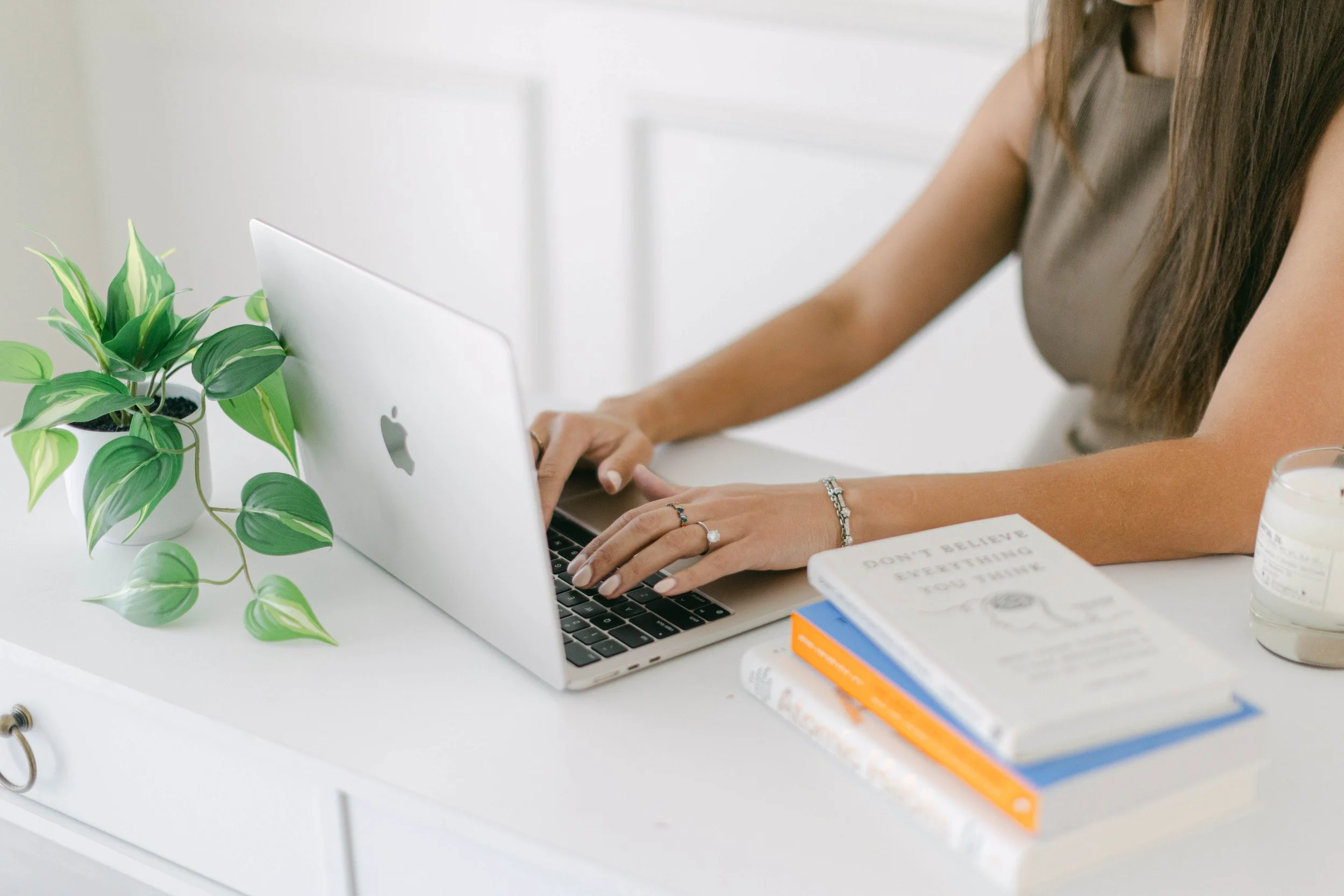 A person with long brown hair working on a silver MacBook laptop at a white desk. The desk has a green potted plant, a white book titled 'Don't Believe Everything You Think,' and some other books. The background is blurred.