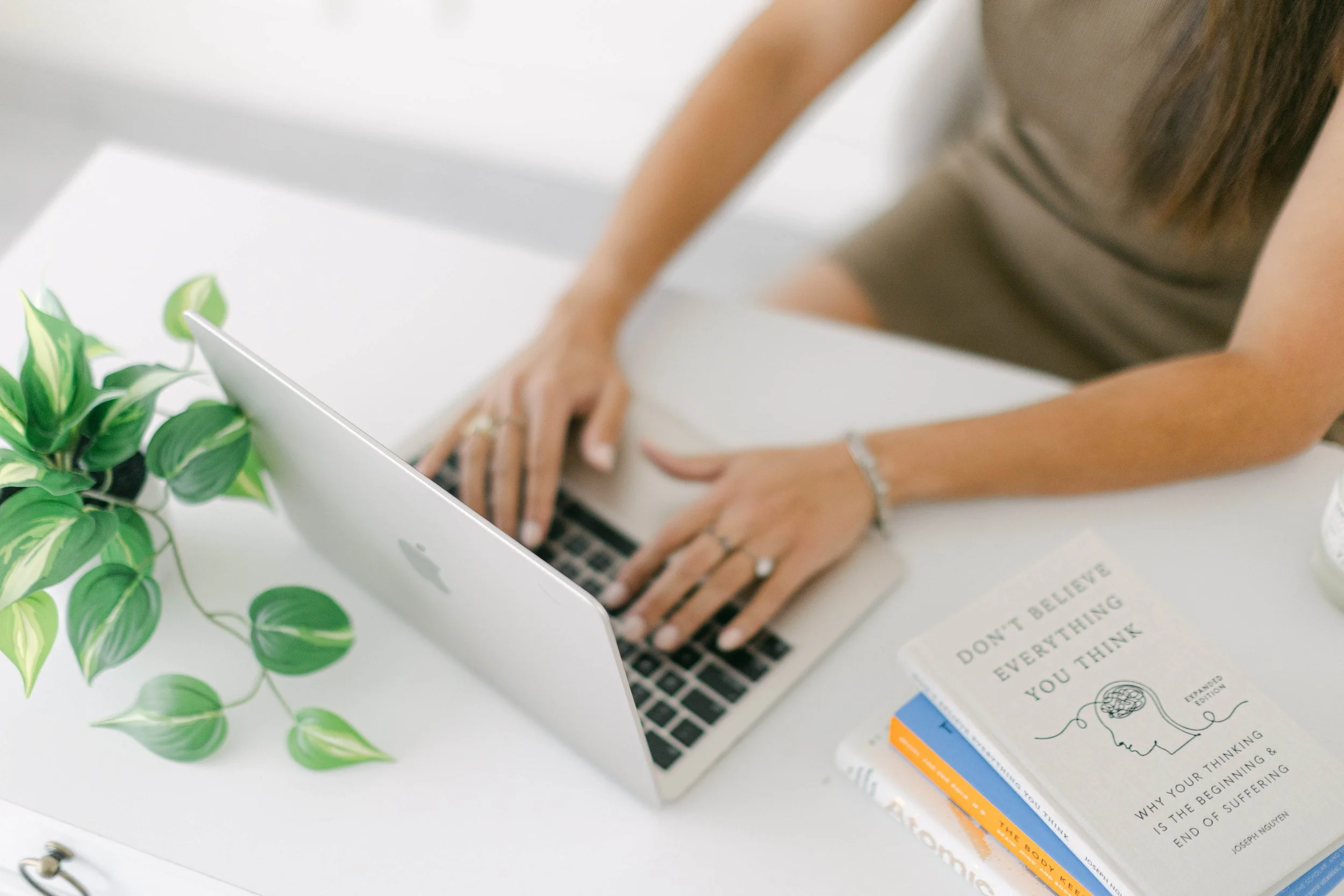 A person sitting at a white desk working on a laptop with a potted plant nearby and a book with motivational quotes.