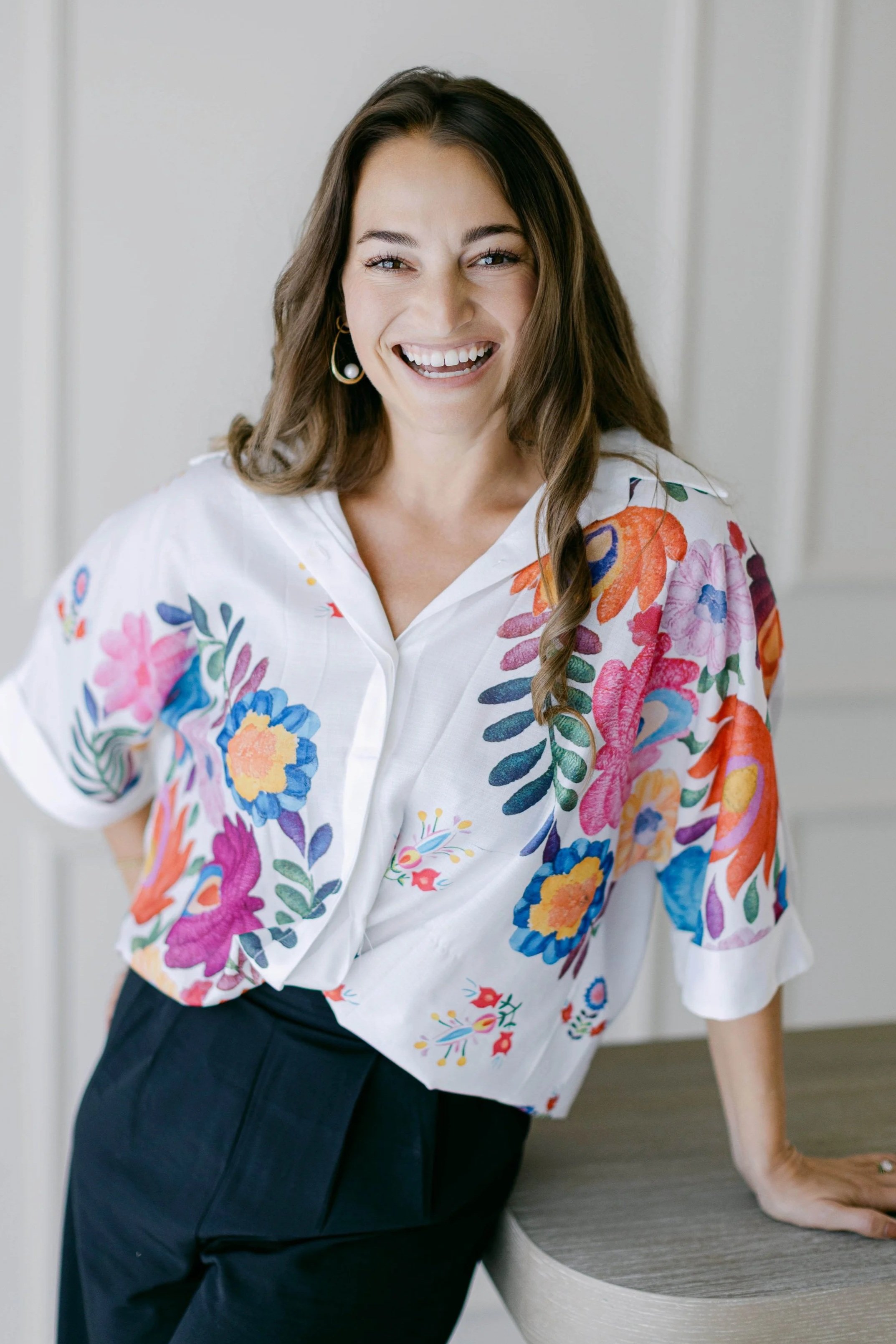 A woman smiling, wearing a white shirt with colorful floral embroidery, black pants, and hoop earrings, leaning against a bathroom countertop.