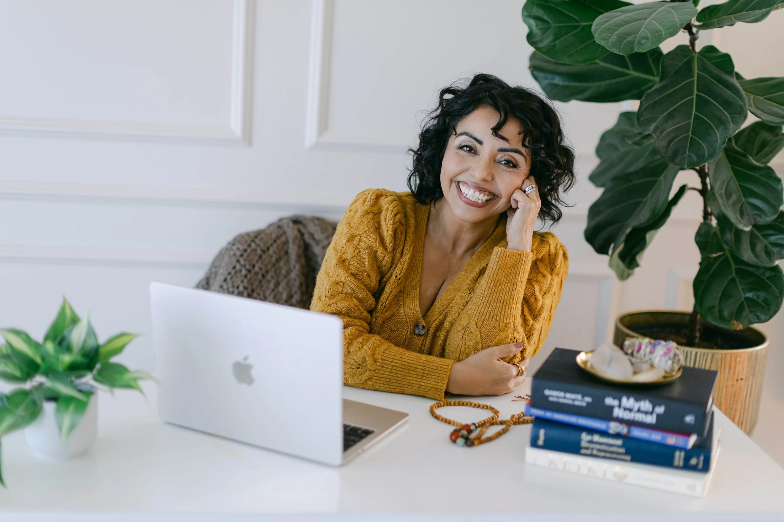 A woman with curly dark hair and a big smile, wearing a mustard yellow sweater, sitting at a white desk with a laptop, a potted plant, and a stack of books in a bright, modern room.