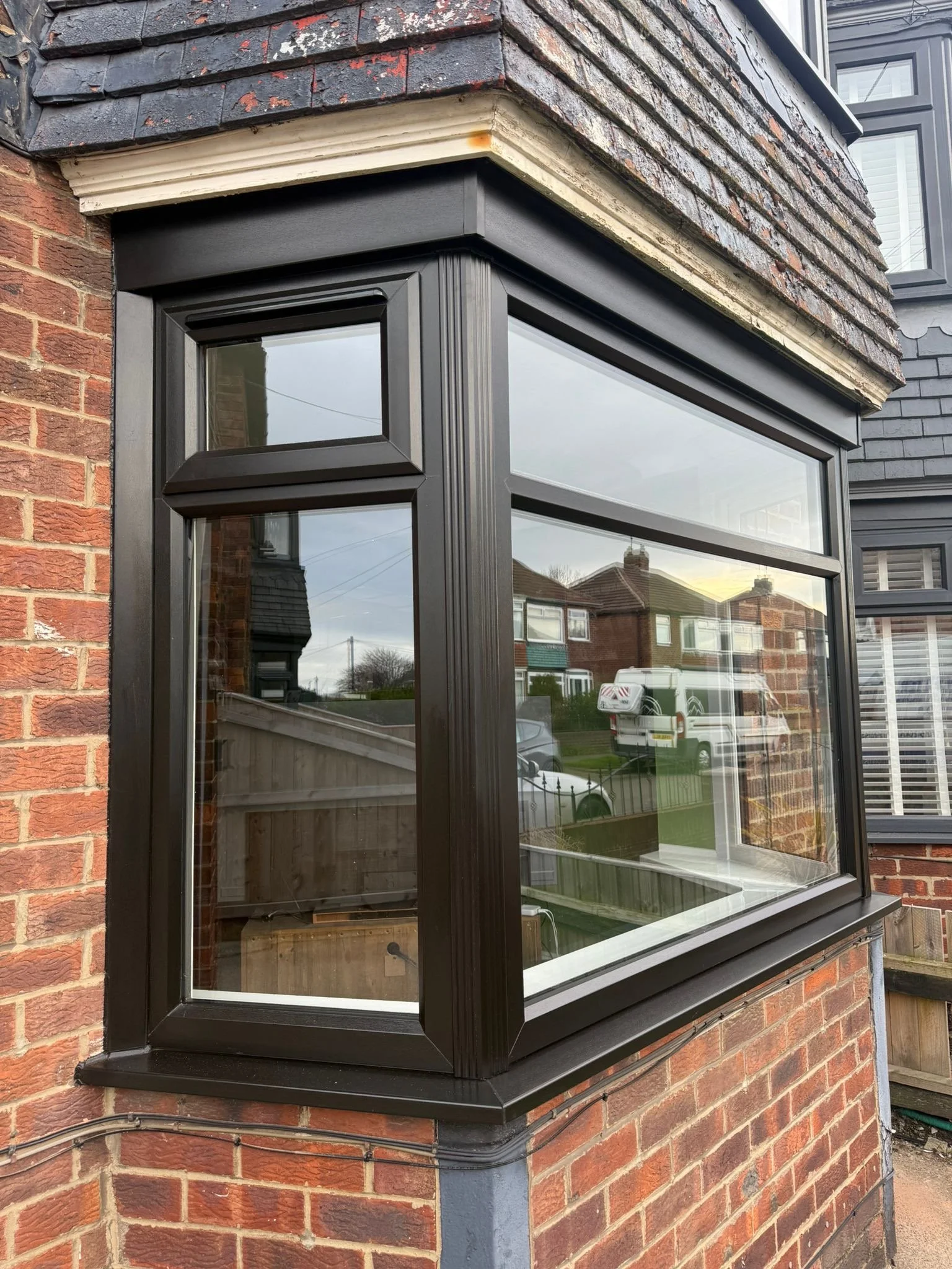 Close-up of a bay window with black frame and glass panes on a brick house with black shingles above.
