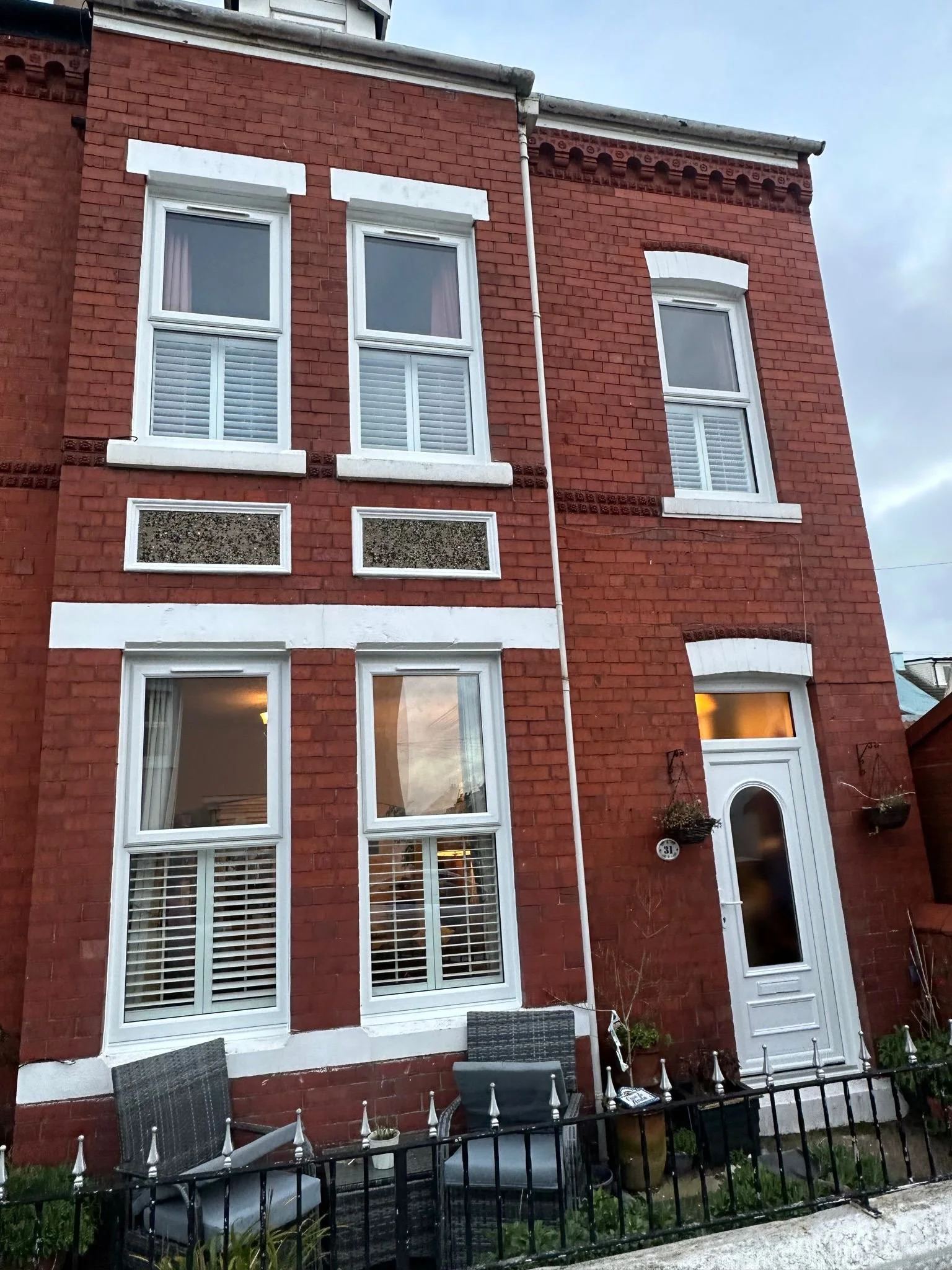 Red brick house with white window frames, a door, and a small fenced patio with chairs and outdoor decorations.