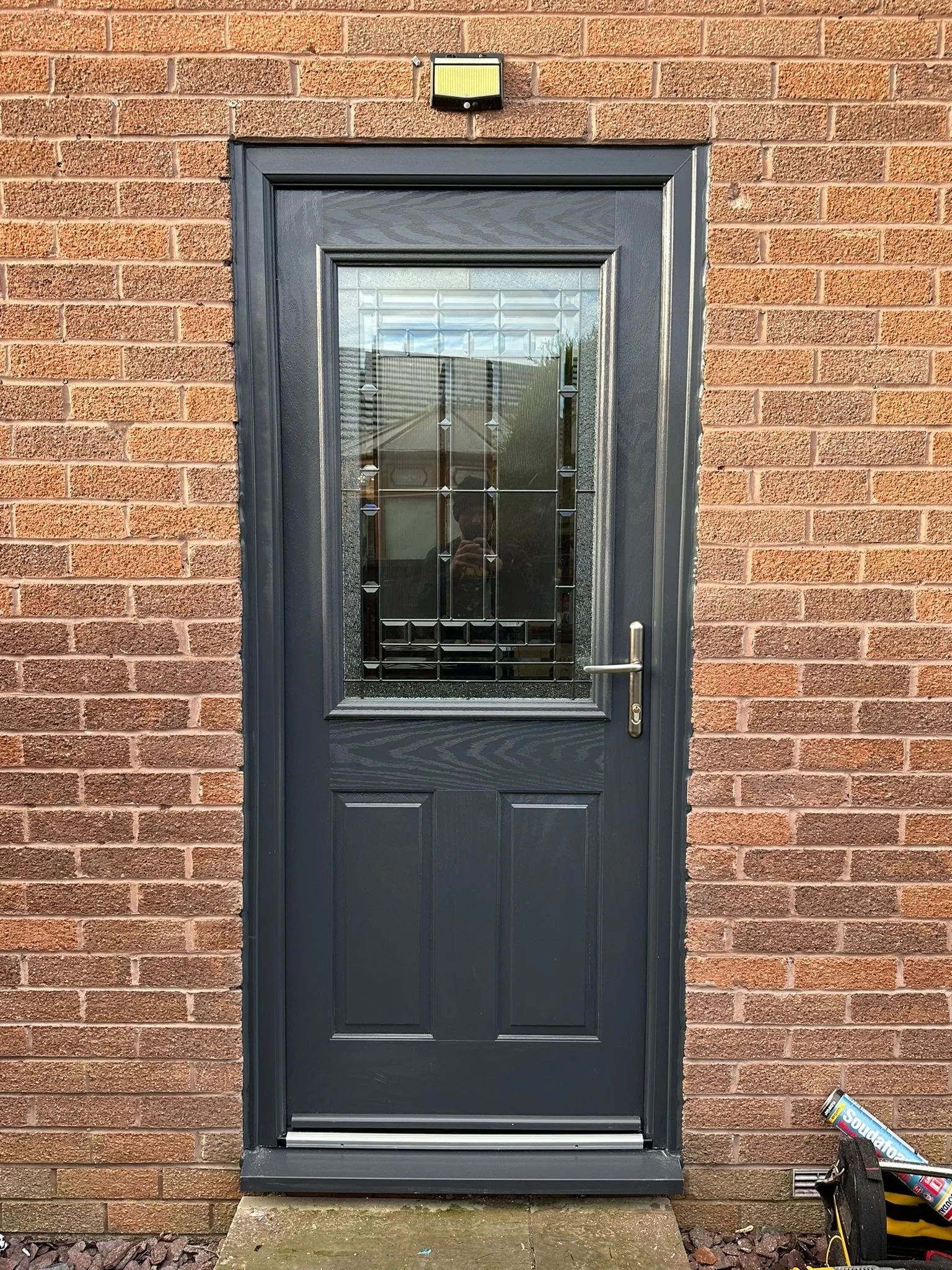 A black door with a decorative glass panel, set into a brick wall. Next to the door on the right, there is a black bag with tools and a box of Dip It spray foam on the ground.