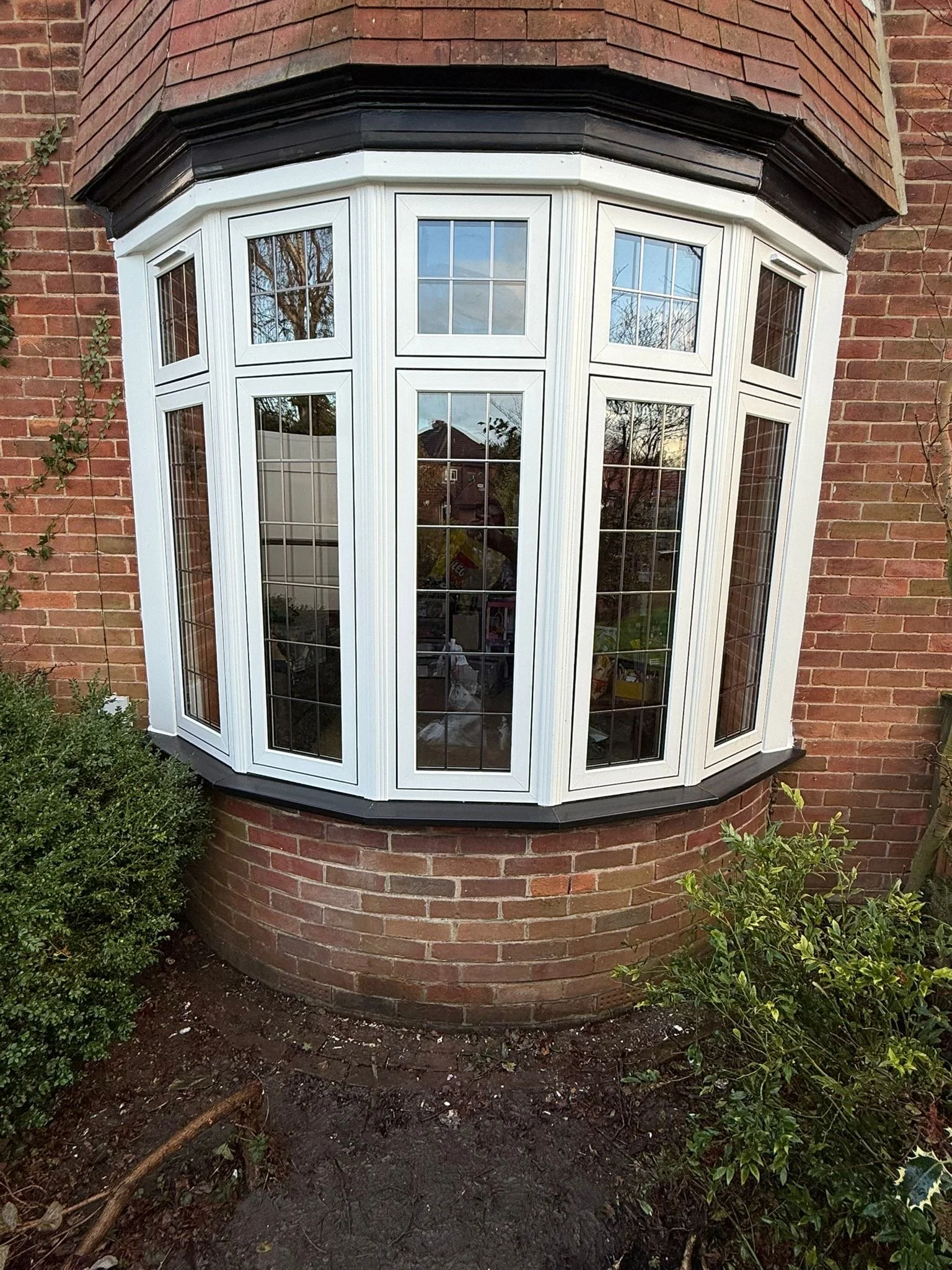 A brick house with a bay window, plants around the foundation, and a patch of soil in front.