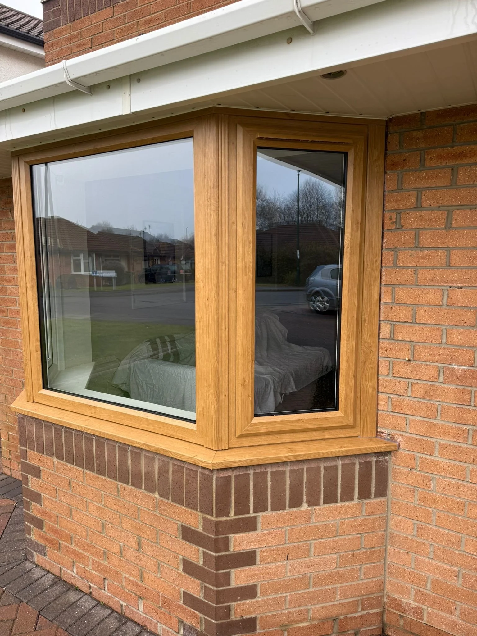 Close-up of a corner window with wooden trim on a brick house, showing the reflection of nearby houses and a street with parked cars.