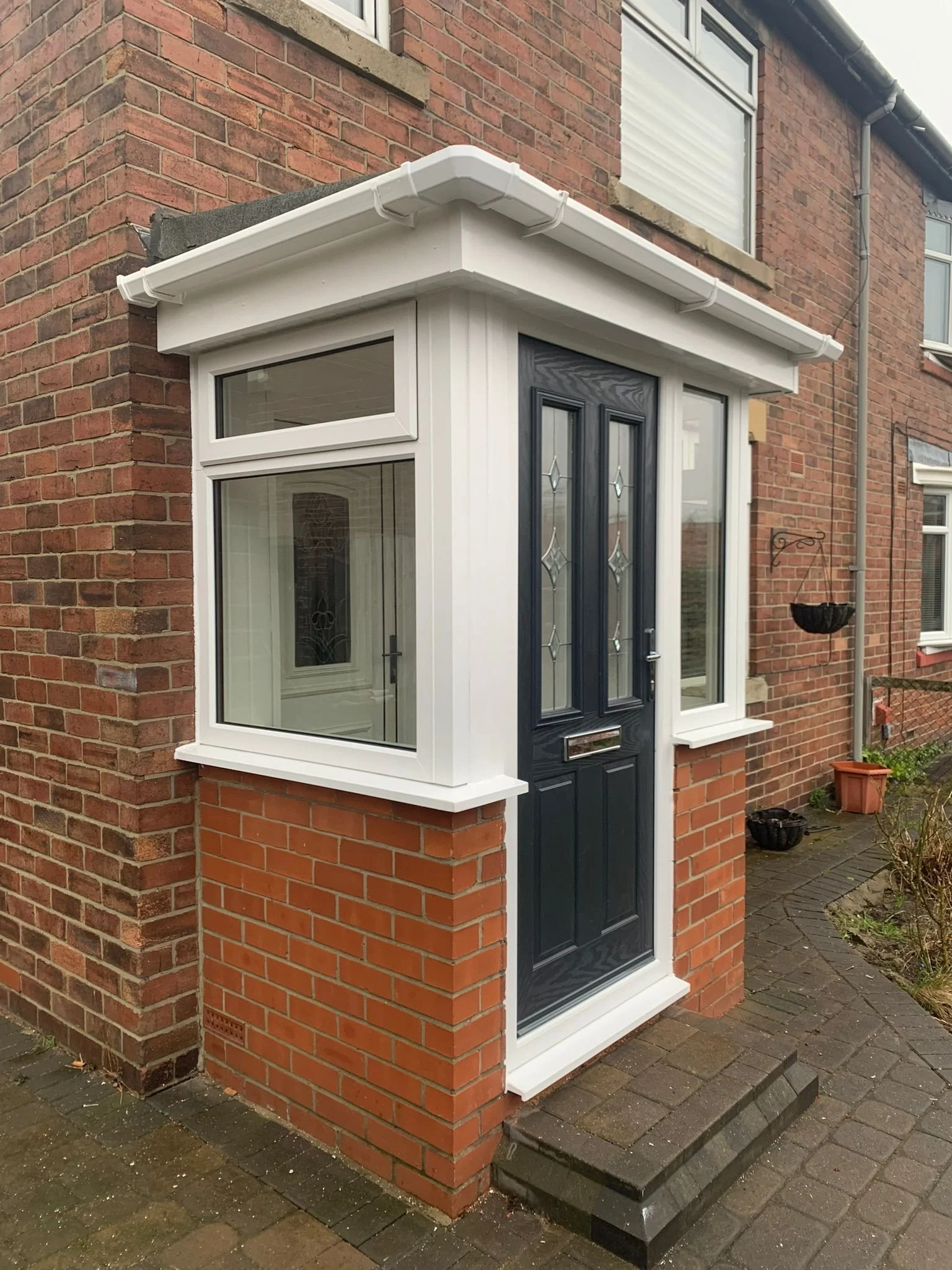 Close-up of a red brick house corner with a white bay window and a black front door with glass panels, small steps, and a concrete paving.