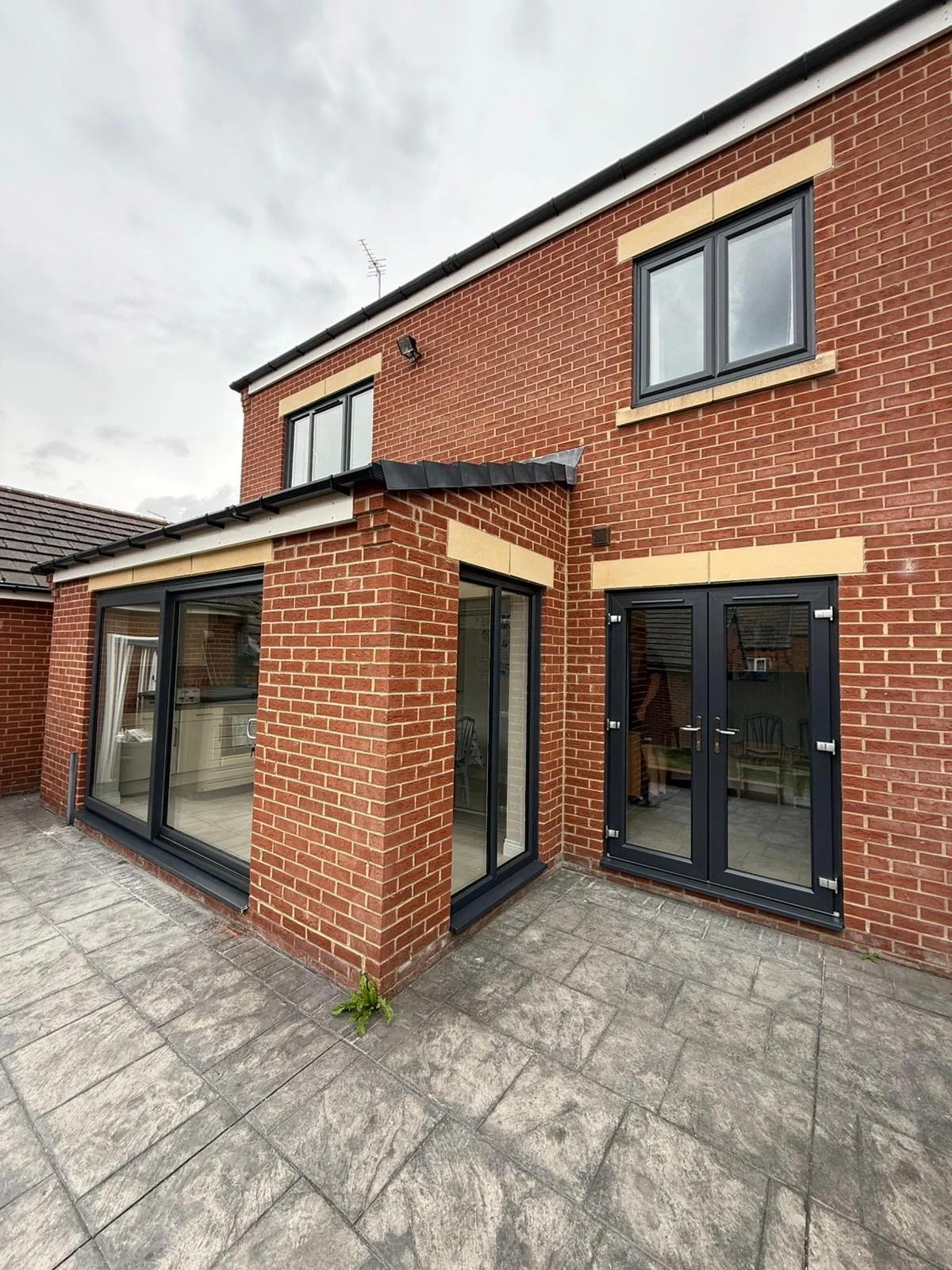 Exterior view of a modern brick house with large black-framed glass doors and windows, a small balcony with a black canopy, and a tiled patio area.