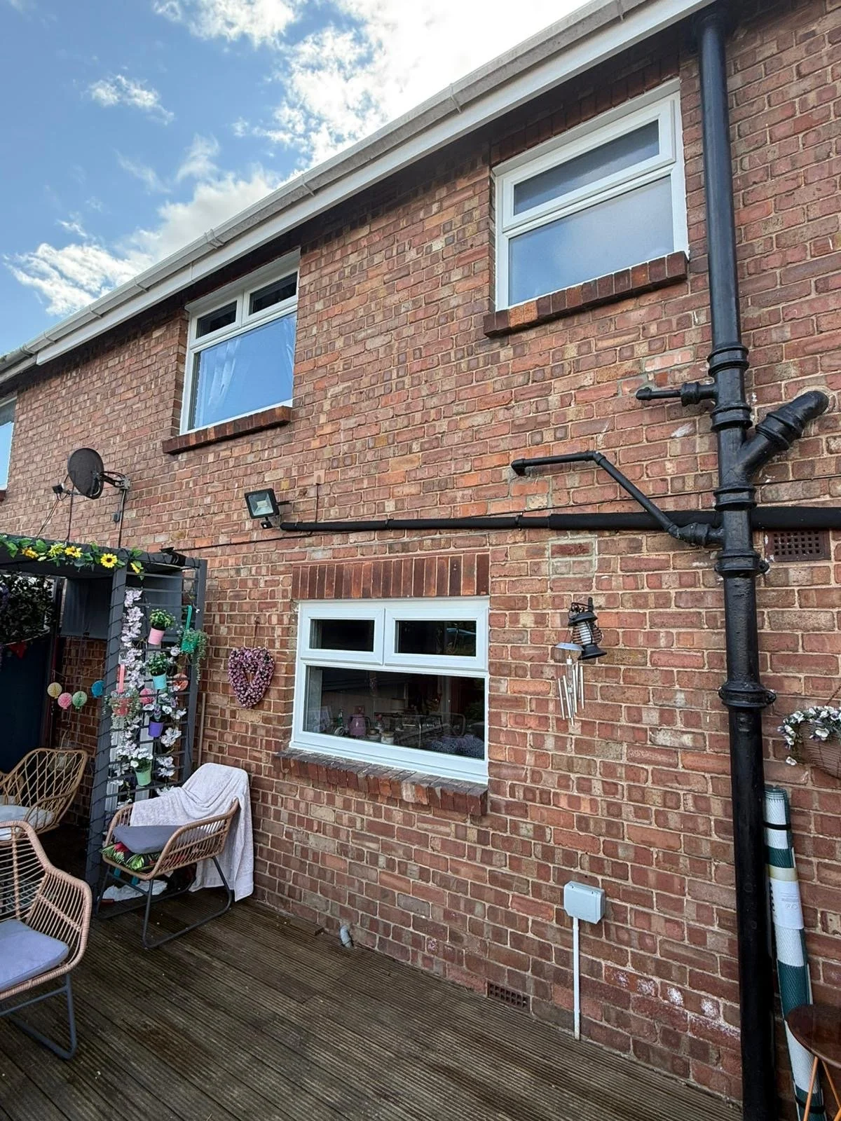 Backyard patio with brick house wall, windows, black drainage pipes, satellite dish, outdoor furniture, flower decorations, and garden ornaments.