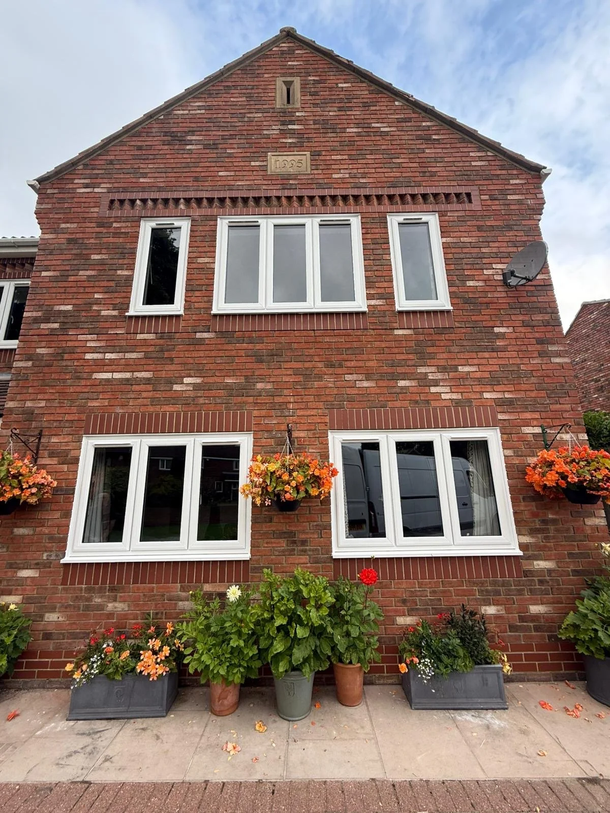 A red brick multi-story house with white-framed windows and hanging flower baskets, and potted flowers on the sidewalk in front.