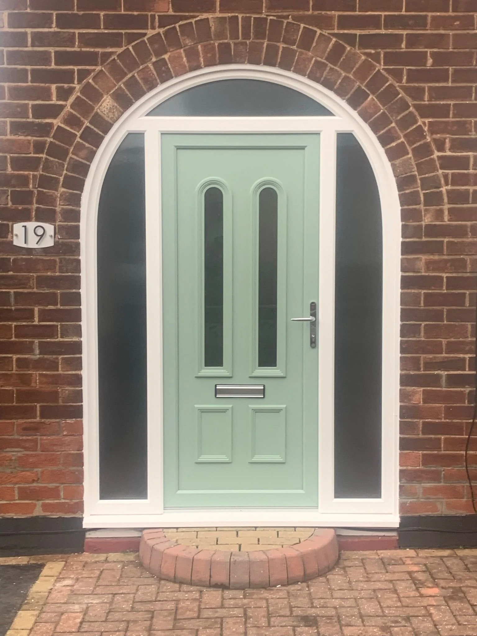 A front door with a light green color, surrounded by a white arched frame and glass side panels, set into a brick wall. The house number '19' is on a small white plaque to the left of the door. A small brick step with a curved front edge is in front 