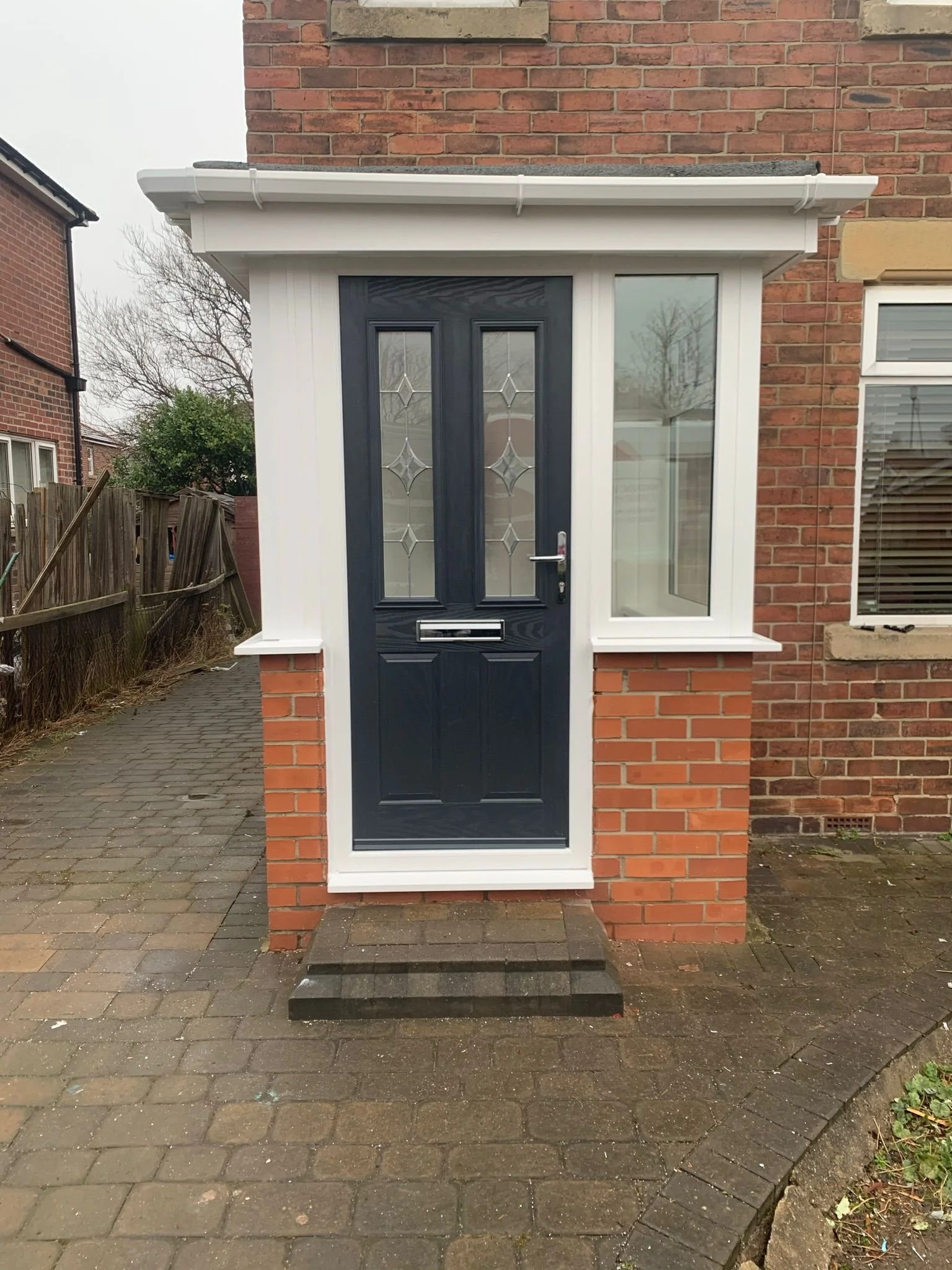 A small porch with a black front door set in a brick wall, flanked by two white frames with glass windows, and raised by a small step on a paved pathway.