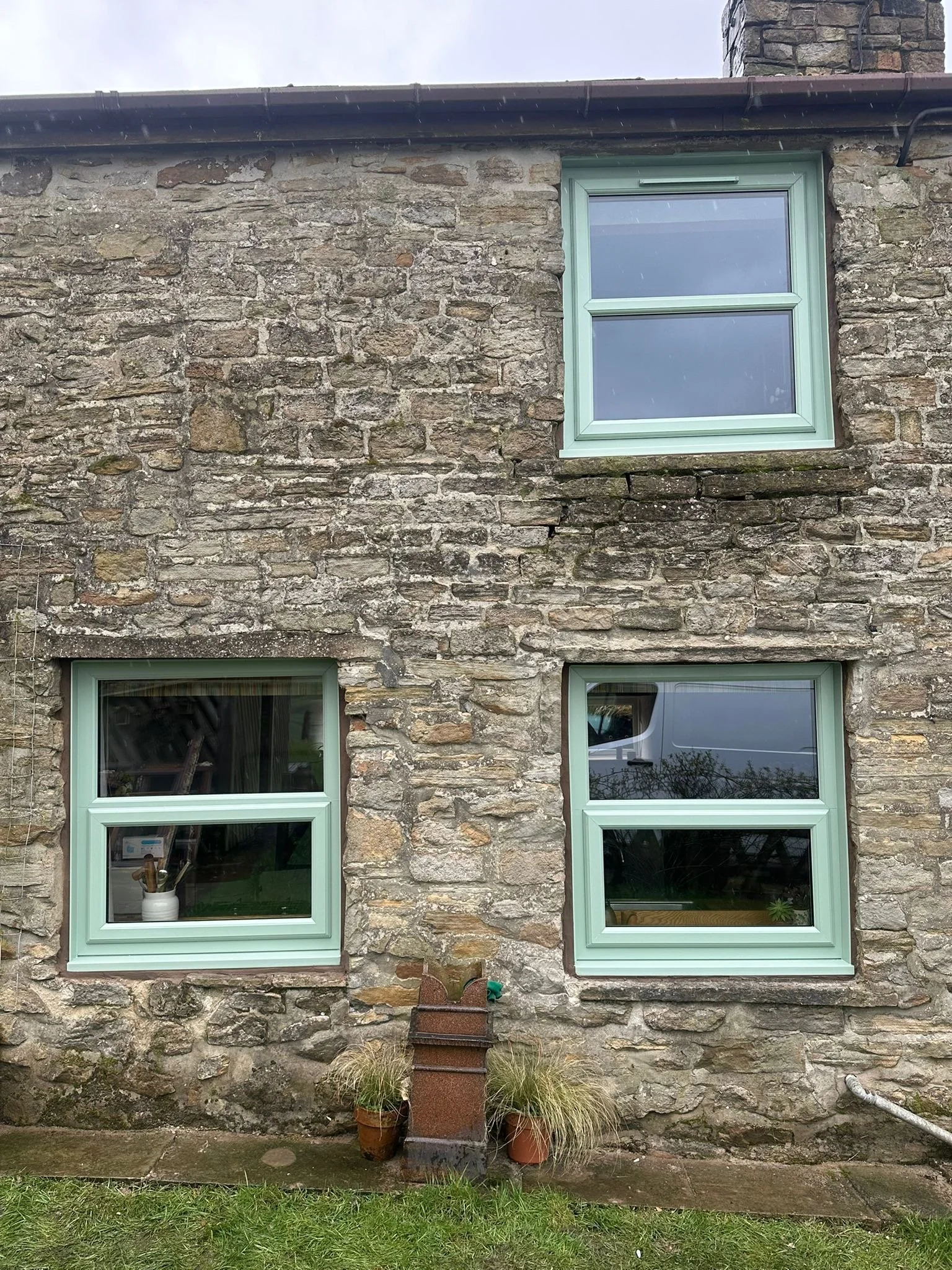 The photo depicts a stone exterior wall of a building with three light green-framed windows. In front of the wall, there are various potted plants and a decorative metal sculpture.