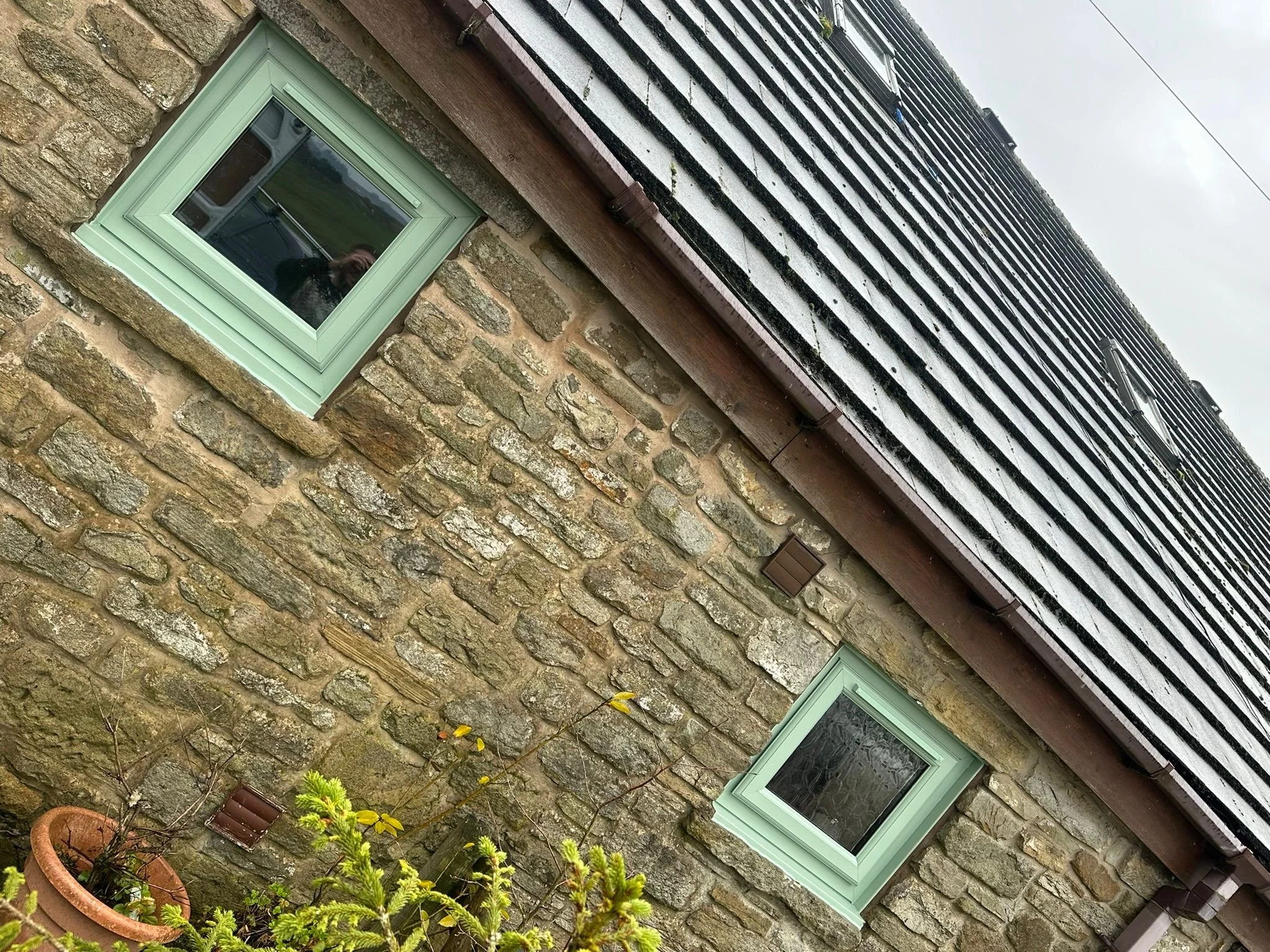 Close-up of a stone house wall with a light green window frame and a black, striped roof with a cable running above.