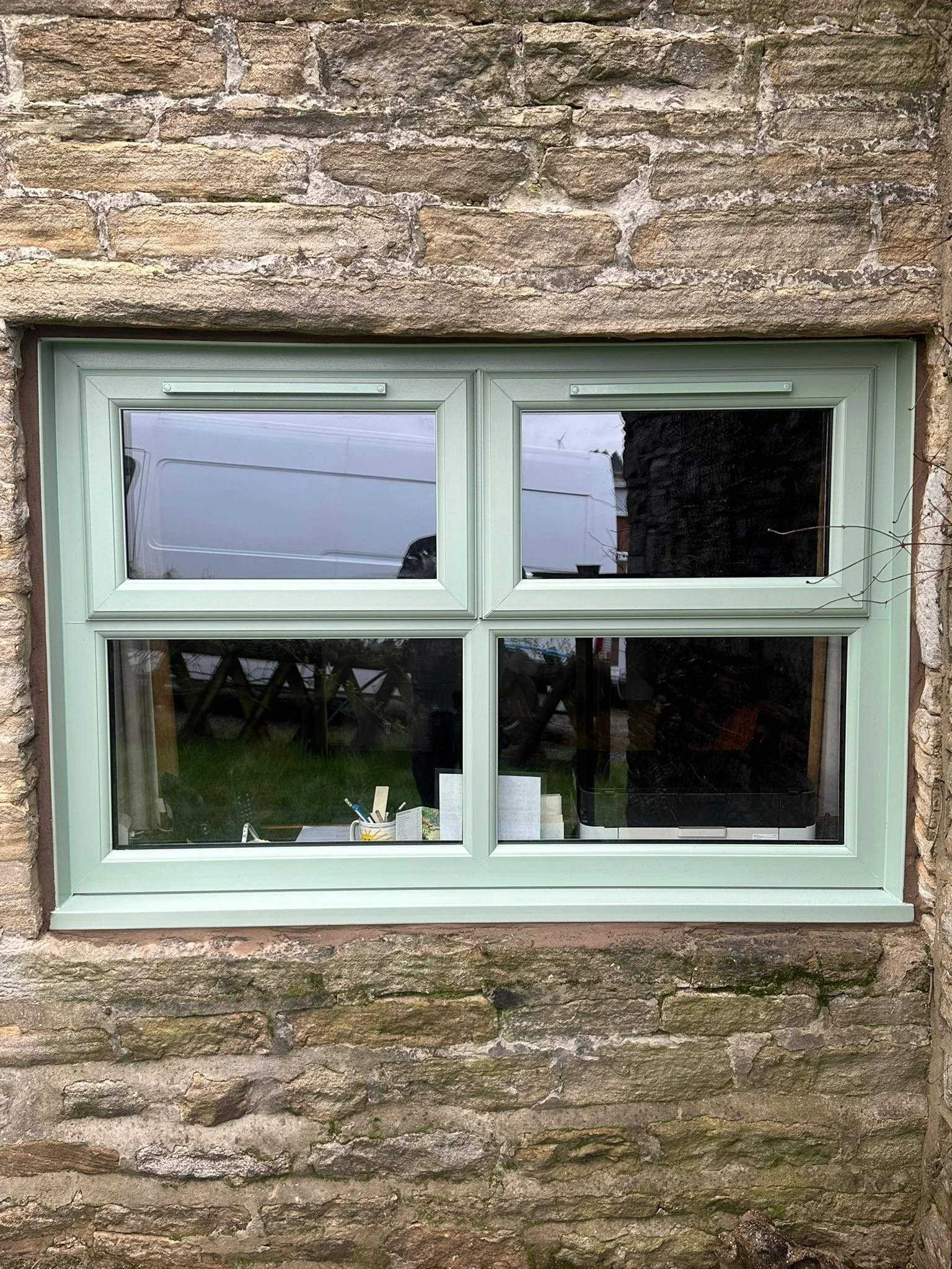 A windowsill with a mint green frame set in a textured stone wall. The window has four panes, with reflections of the outdoor scene visible on the glass.
