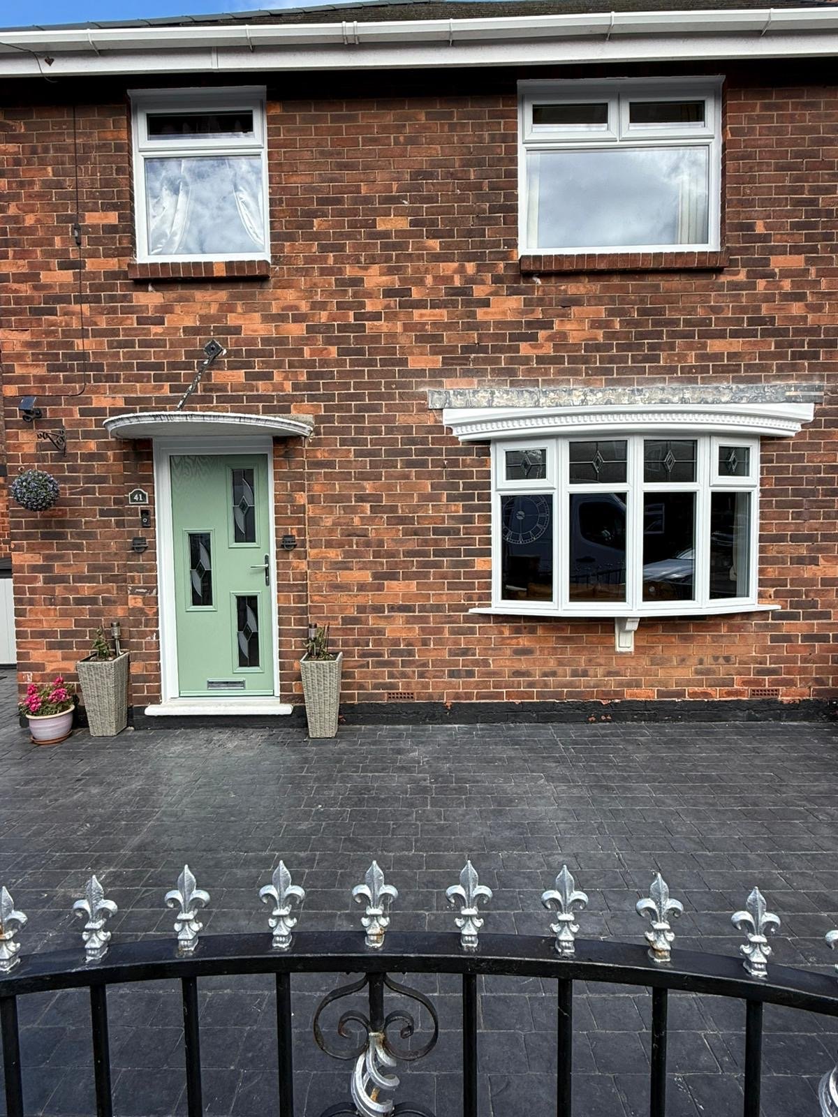 Front of a brick house with a green door, two potted plants, a large white bay window, and two upper windows. The house has a black fence with silver decorative elements.