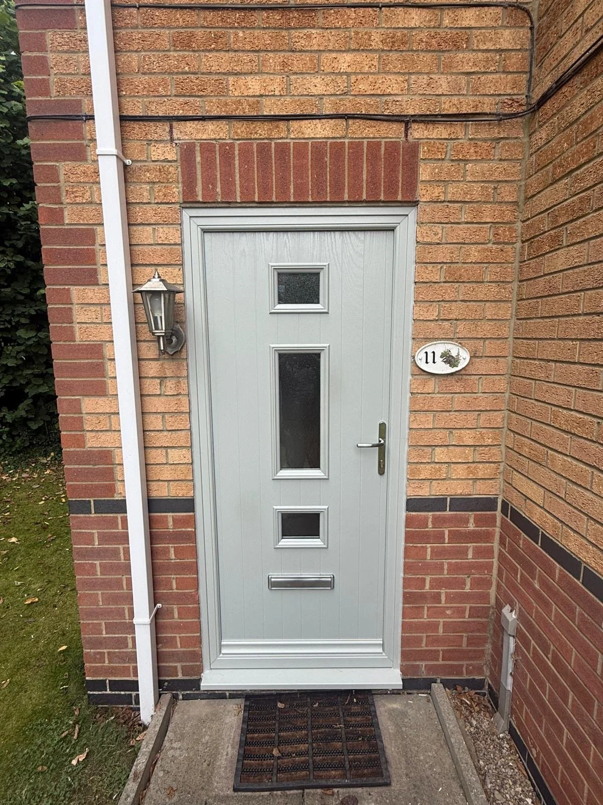 A white front door with rectangular glass panes on a brick wall. To the left of the door is a wall-mounted lantern-style light. A house number sign with the number 11 and a decorative flower is on the wall to the right of the door. A black mat with a