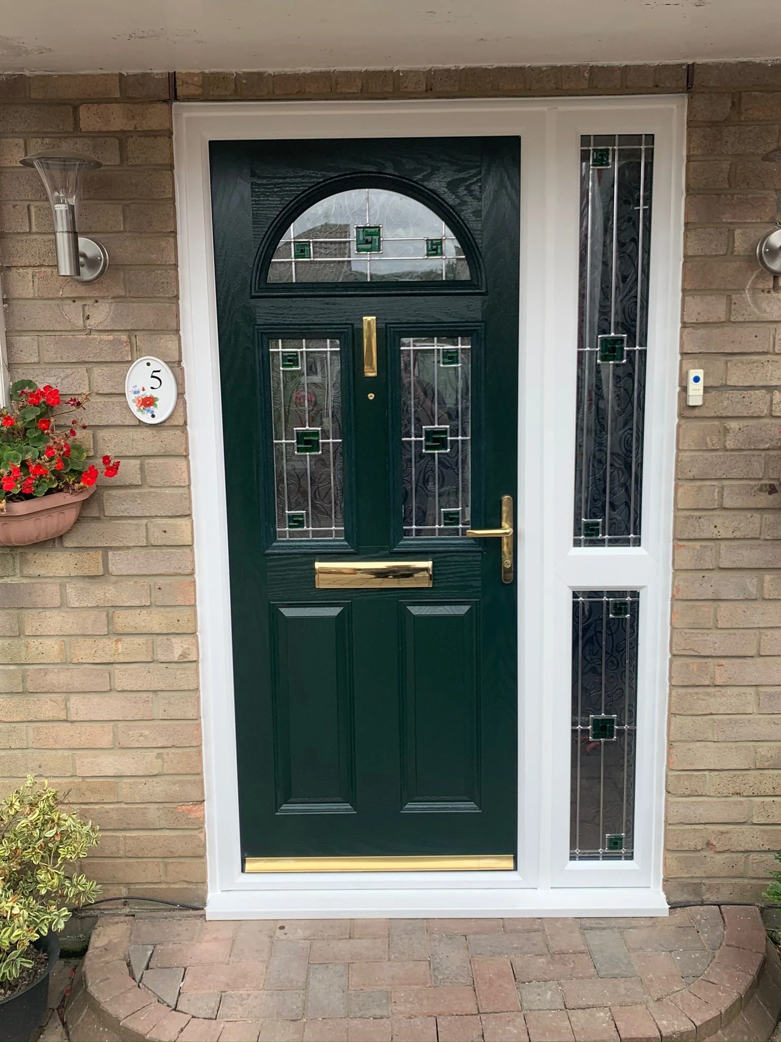 Front door with dark green color and a decorative glass window above and to the side, set in a brick wall entrance with potted plants and a house number sign with the number 5.