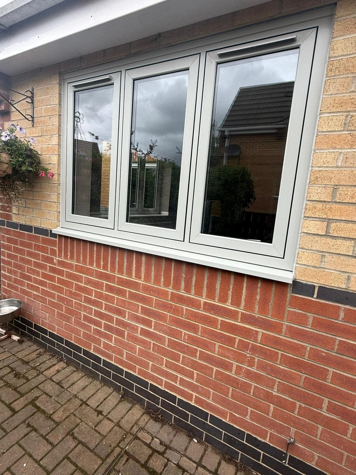 A brick house exterior featuring a triple-pane white-framed window with a reflection of the sky and nearby houses. There is a concrete flower box with flowers on the left and a small metal pet dish on the ground in the bottom left corner. The base of