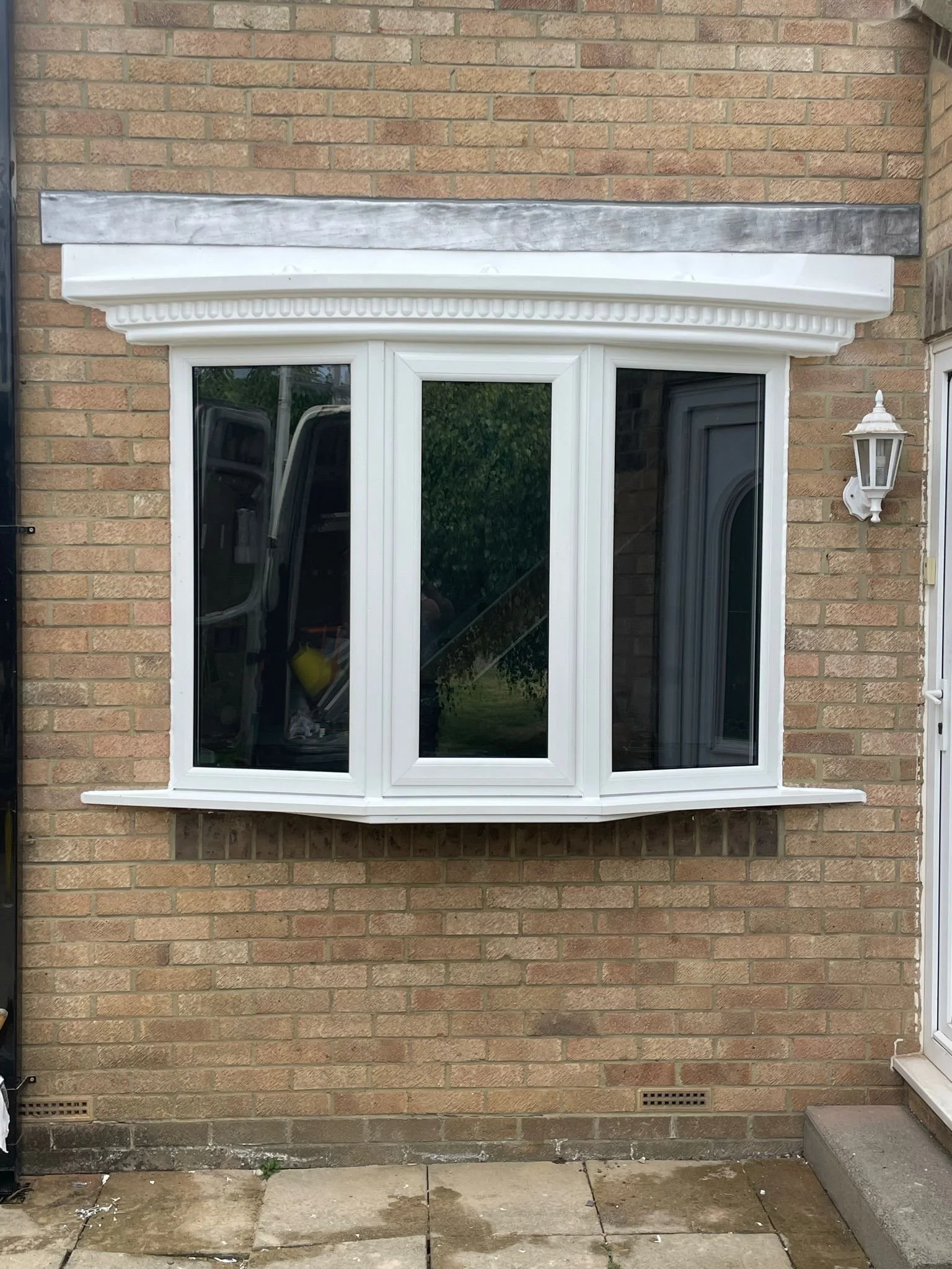 A white bay window with decorative molding on an exterior brick wall. To the right of the window, there is a white wall-mounted outdoor lantern. The ground below consists of concrete patio stones.
