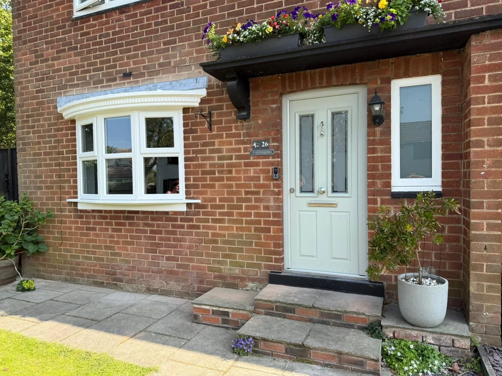 Brick house entrance with white door, two windows, flower boxes with purple, yellow, and white flowers, and potted plant on the right side