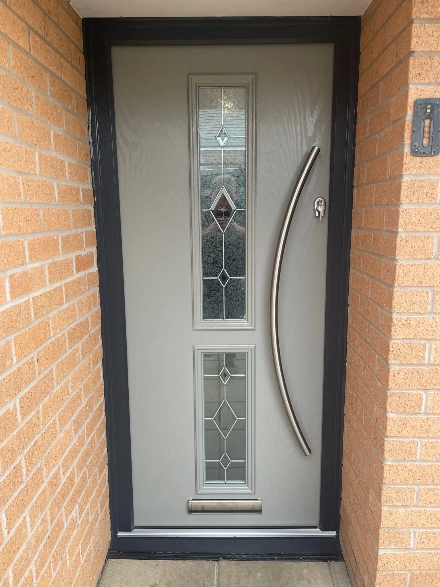 Front door with glass panel featuring decorative diamond shapes, black outer frame, stainless steel handle, curved metal entry bar, and brick walls on either side.